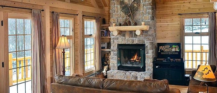 Cozy log cabin living room with stone fireplace, leather sofa, and large windows overlooking snowy outdoors