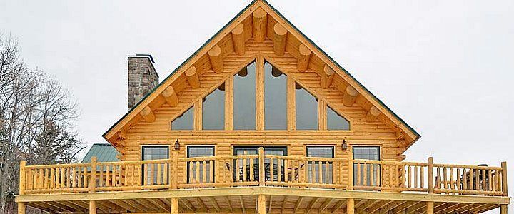 Wooden cabin with a large peaked roof and wraparound deck under a cloudy sky