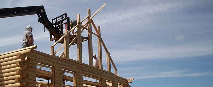 Wooden house frame under construction against a blue sky, with a crane lifting beams.