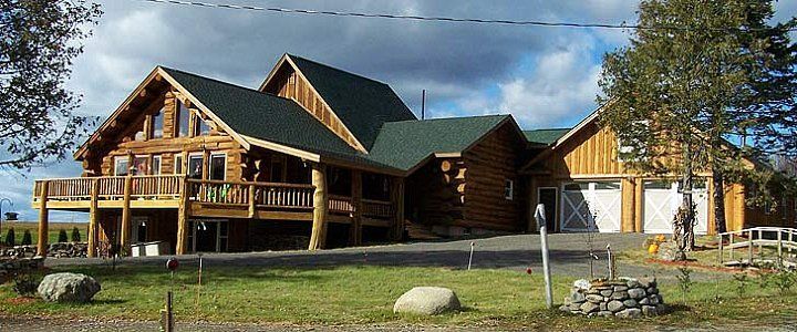 Large rustic log cabin with a green roof, porch, and garage in a wooded yard under a blue sky
