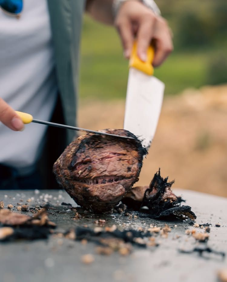 A person uses a knife to carve a charred piece of steak on a metal surface outdoors.