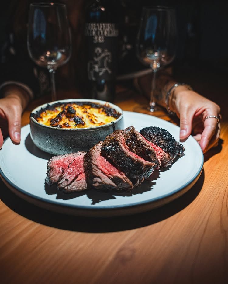 A person holds a plate with sliced steak and a side of gratin, with wine glasses and a bottle on a wooden table.