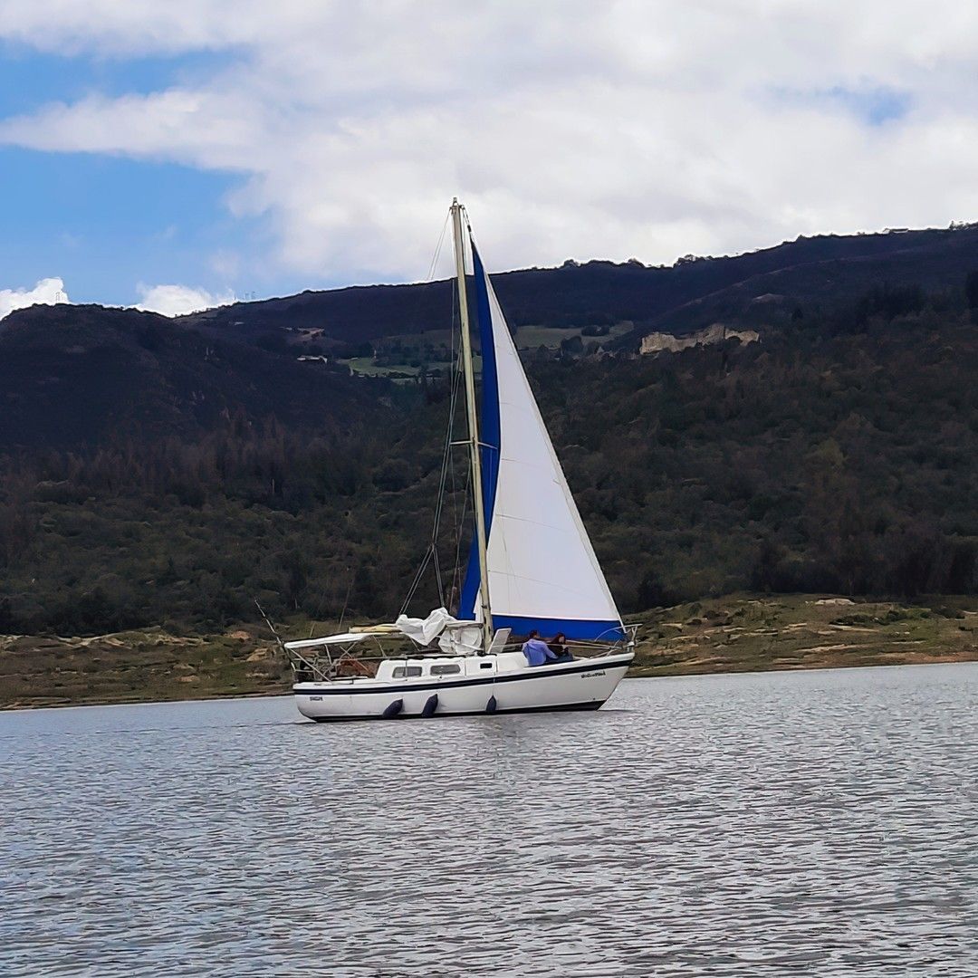 A white sailboat with a blue and white sail cruising on a body of water with hills in the background.