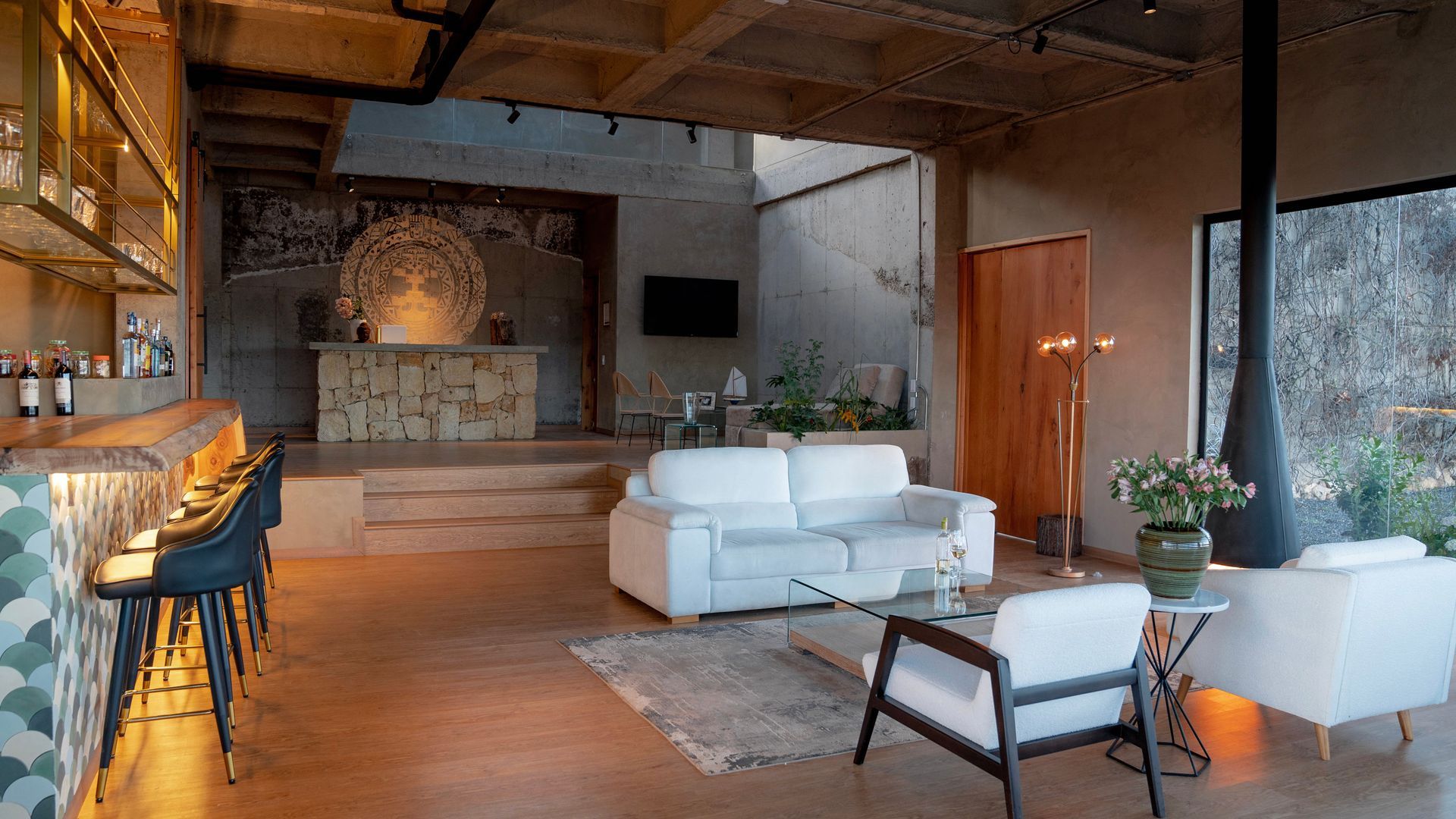 A modern open-plan living room with white sofas, a bar area, and a hanging fireplace in a room with concrete ceilings.