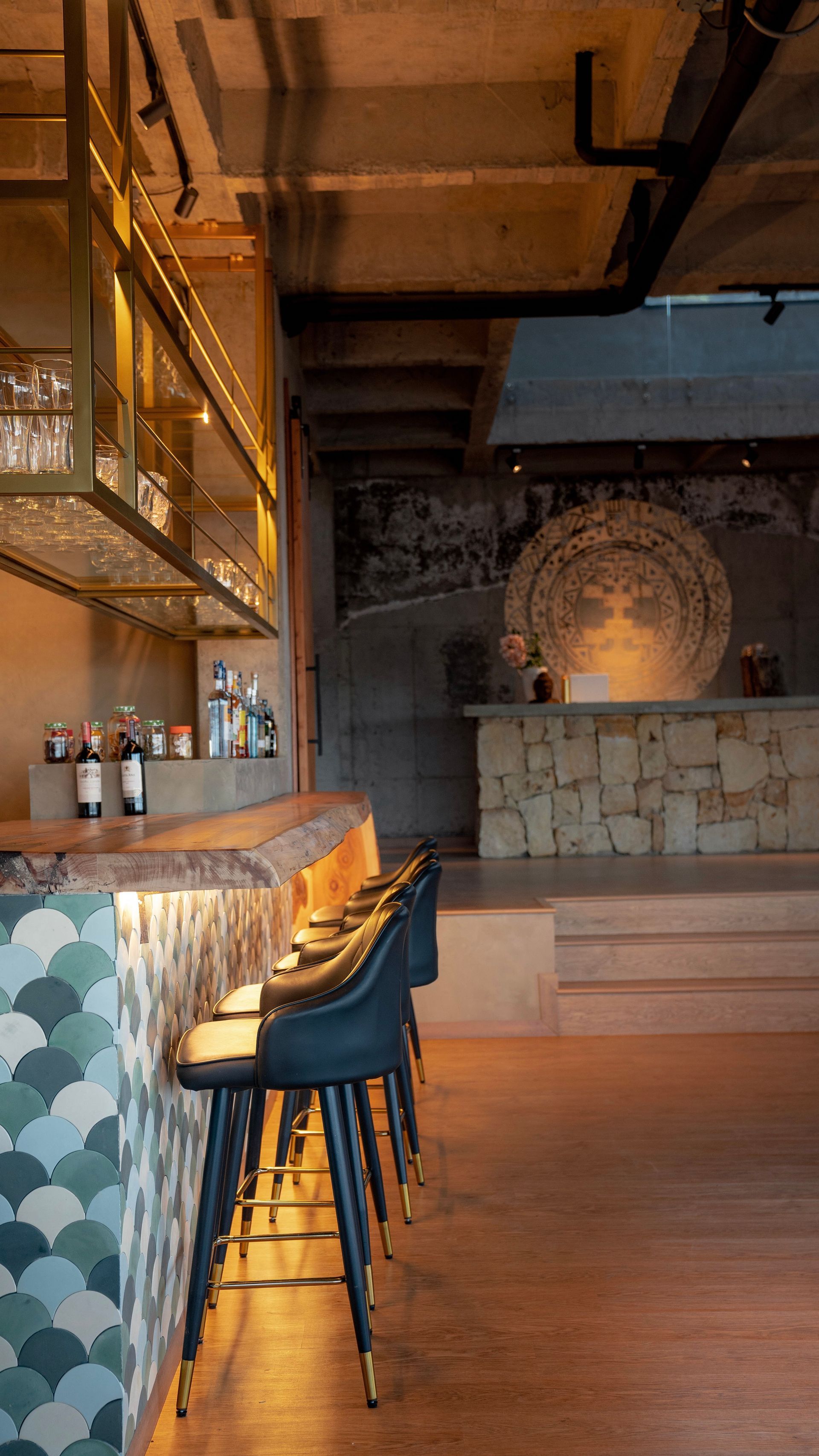 A bar interior featuring a teal scallop-tiled counter with dark stools, a live-edge wood top, and a stone-walled stage area.