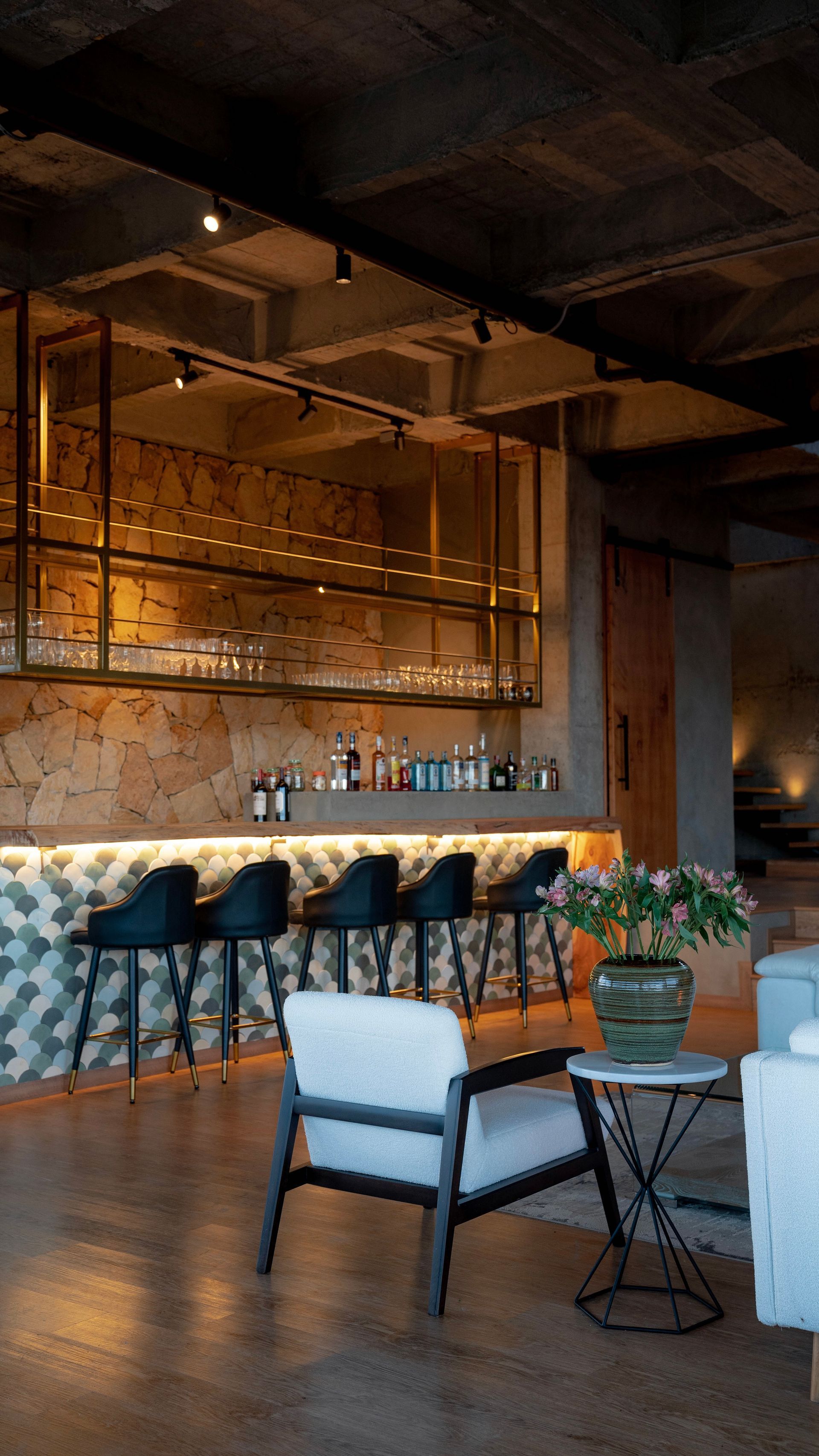 A dimly lit bar with a patterned counter, black stools, and a metal shelf, featuring a white armchair and side table.
