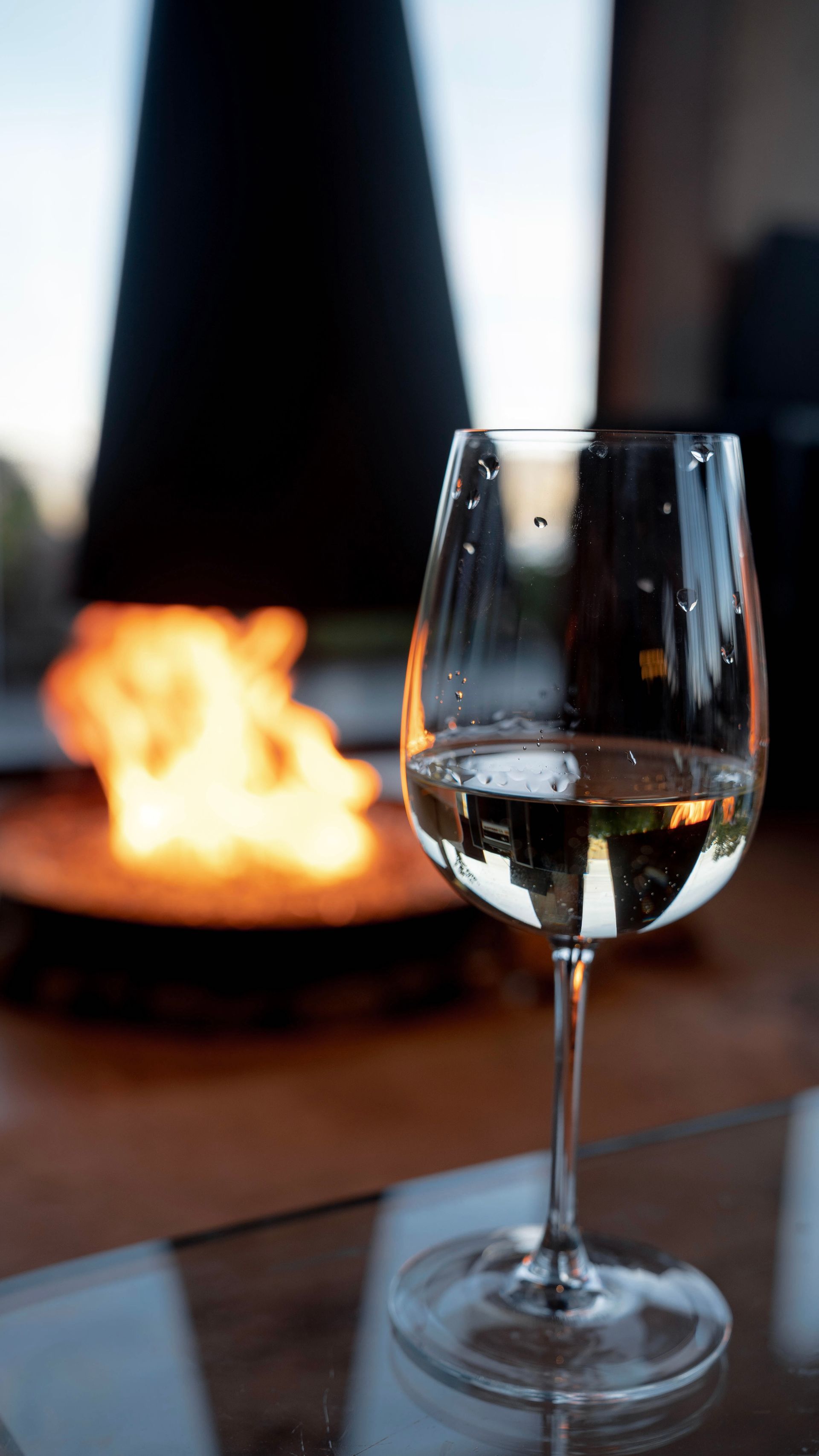 A glass of white wine sits on a glass table in front of a warm fireplace.