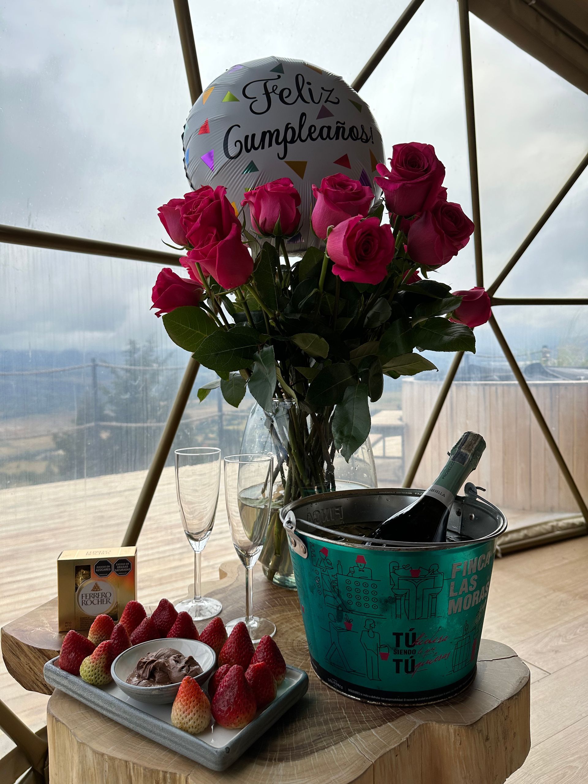 A birthday setup with a bouquet of pink roses, champagne, and strawberries on a wooden table inside a geometric dome.