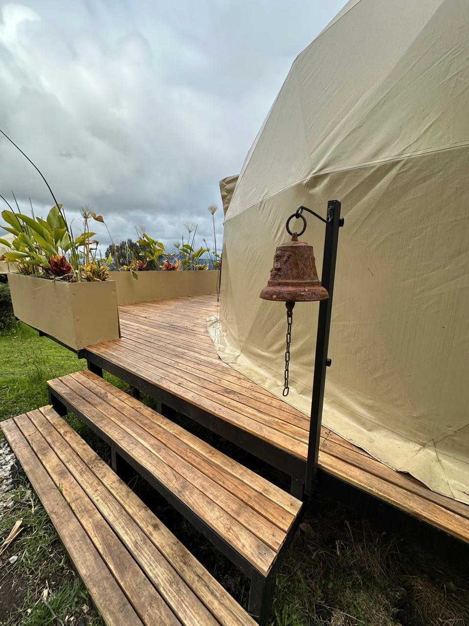 Wooden stairs lead to a deck featuring a tan geodesic dome tent, decorative planters, and a small hanging metal bell.