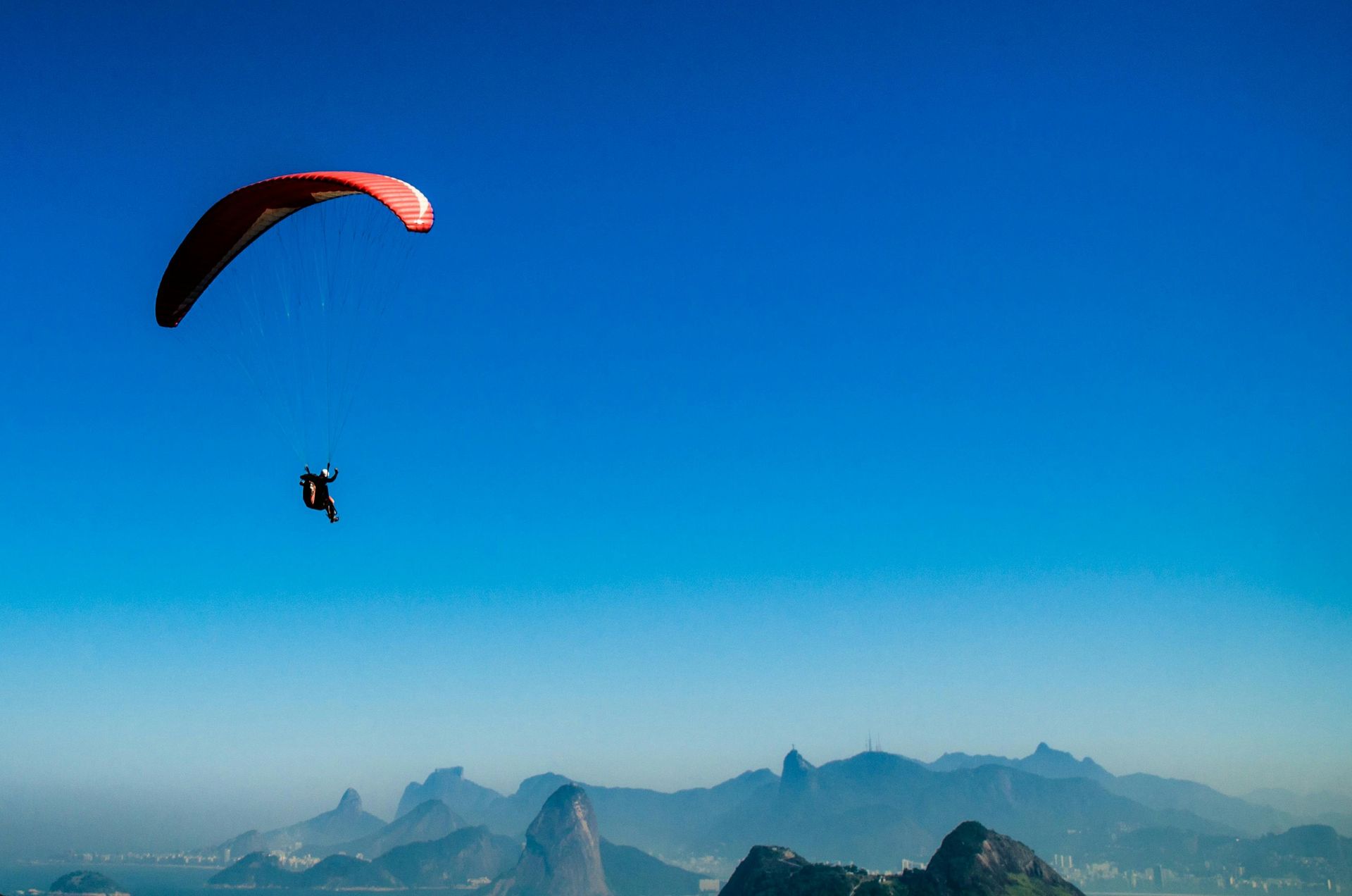 A paraglider with a red wing flies against a clear blue sky over mountainous terrain.