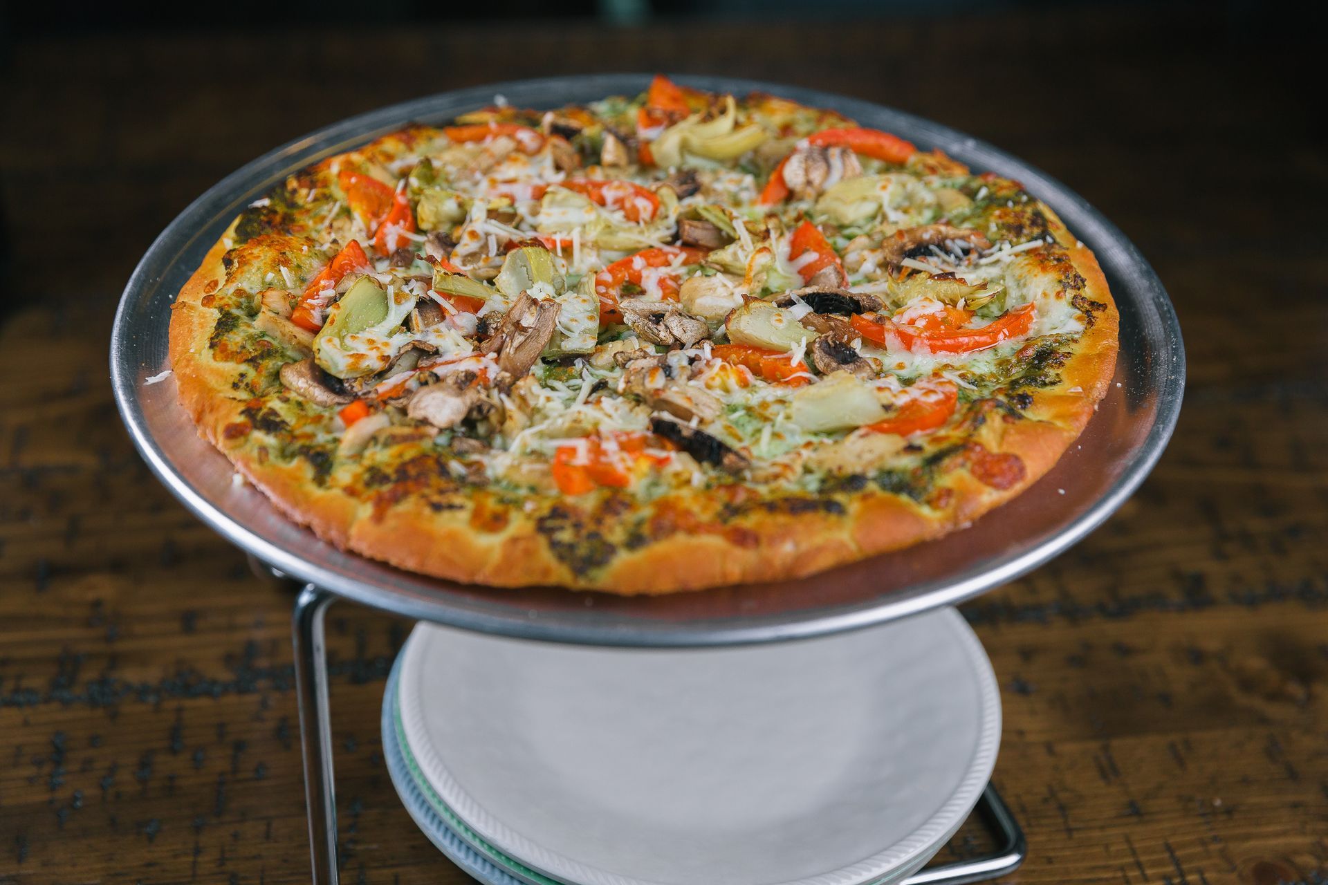 Large vegetable pizza on a metal stand over stacked plates on a dark wooden table