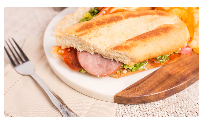 Sandwich on a white plate with lettuce and ham, served with a fork on a light table