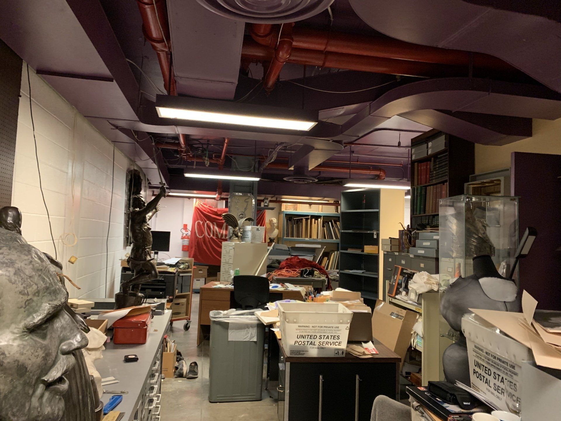 Cluttered basement workspace with desks, storage, and art supplies under purple ceiling fixtures.