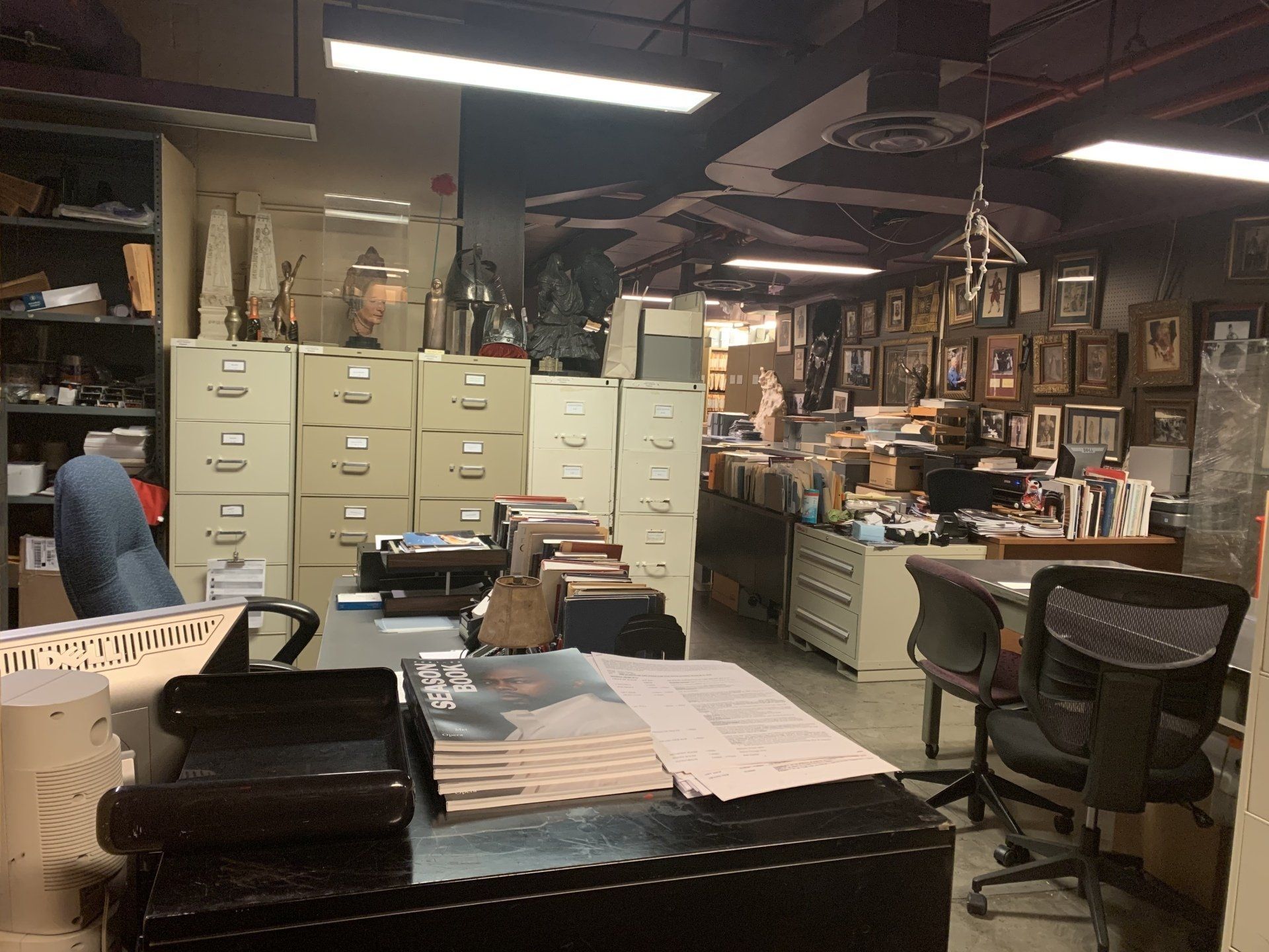 An office filled with filing cabinets, a desk, and various items, with a cluttered, busy atmosphere.