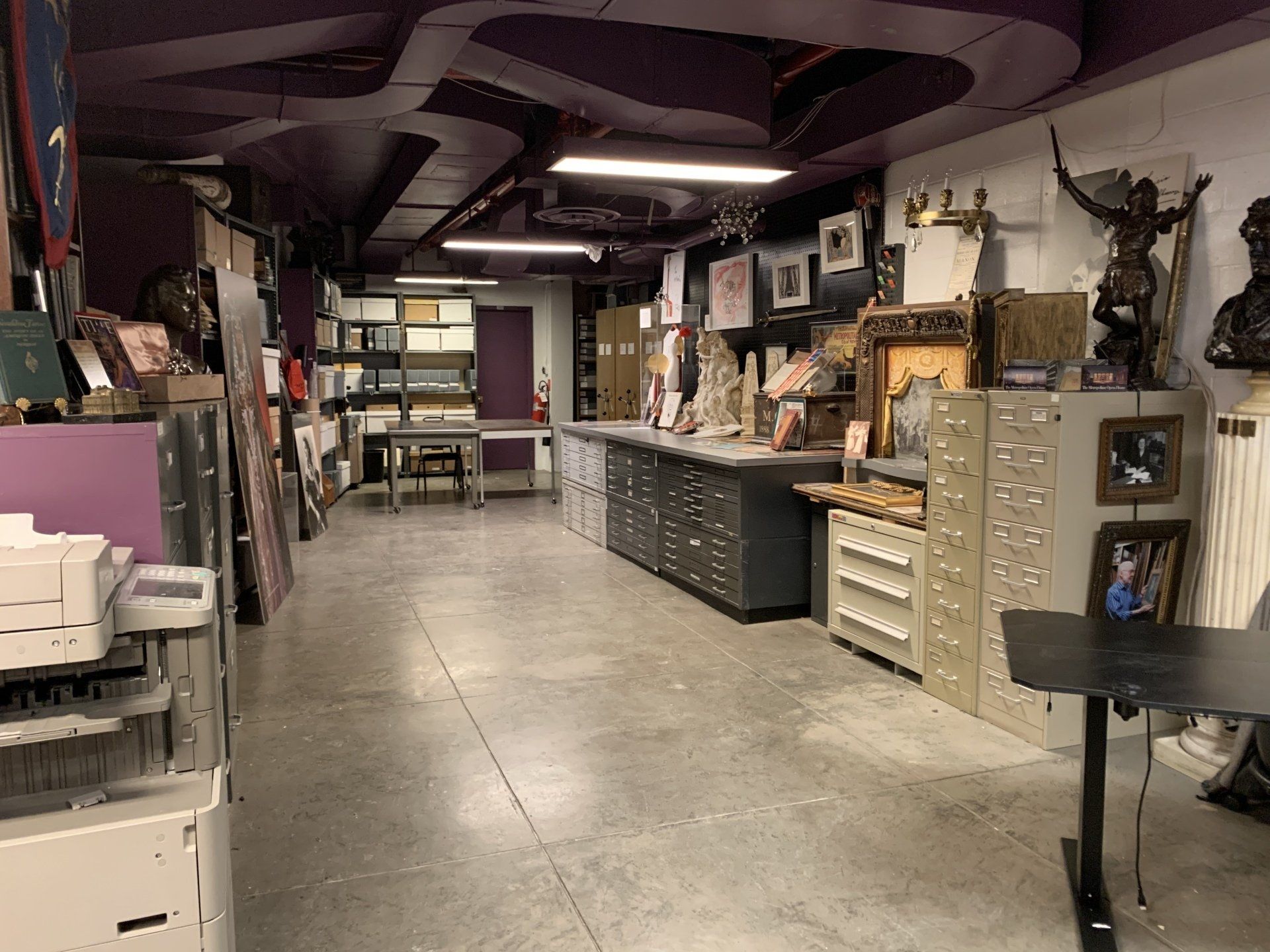 Interior of a cluttered room with art supplies, filing cabinets, and various sculptures. Purple ceiling.