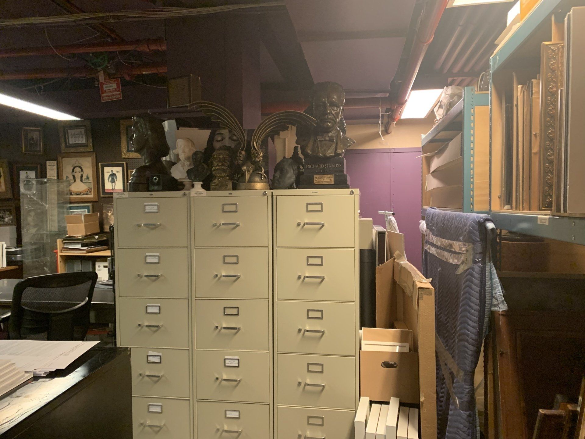 A cluttered storage room with filing cabinets topped with statues and shelves holding artwork.