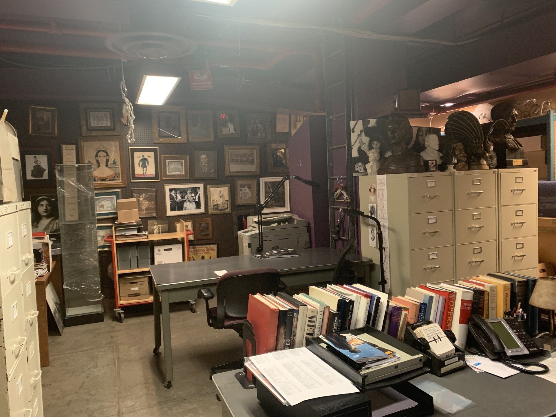 Cluttered office with a desk, filing cabinets, framed photos on the wall, and various objects.