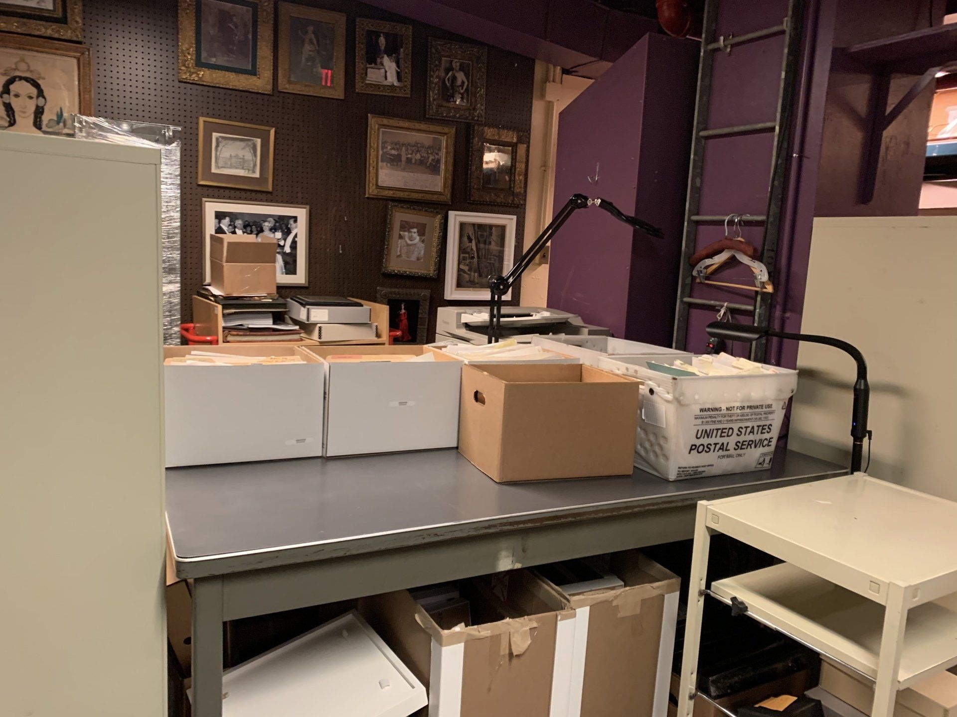 Desk with boxes, photos on wall, lamp, and a small table in a cluttered workspace.
