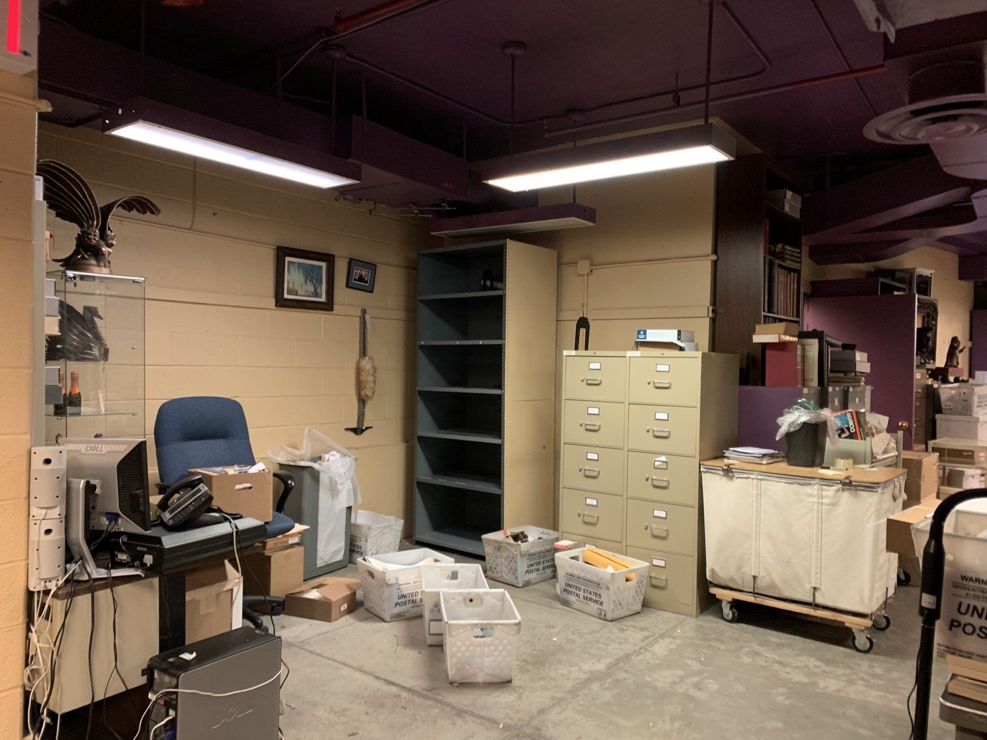 A cluttered office with boxes, filing cabinets, and shelves. Purple ceiling, beige walls.