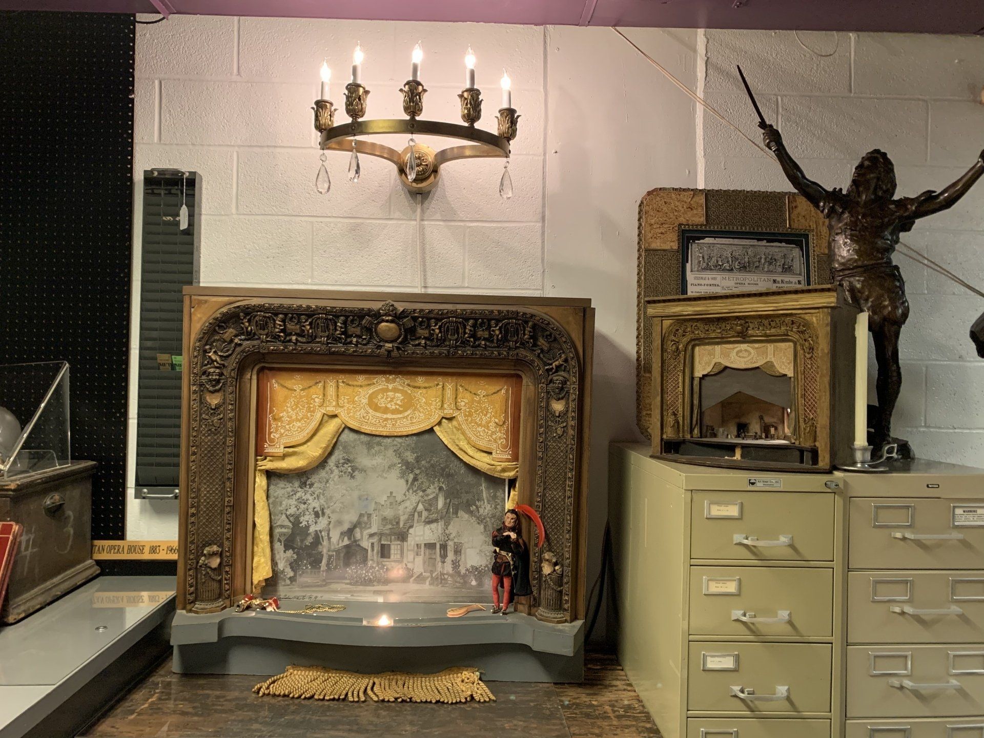 A vintage stage prop with detailed frame, figures, and a brass chandelier against a white wall.