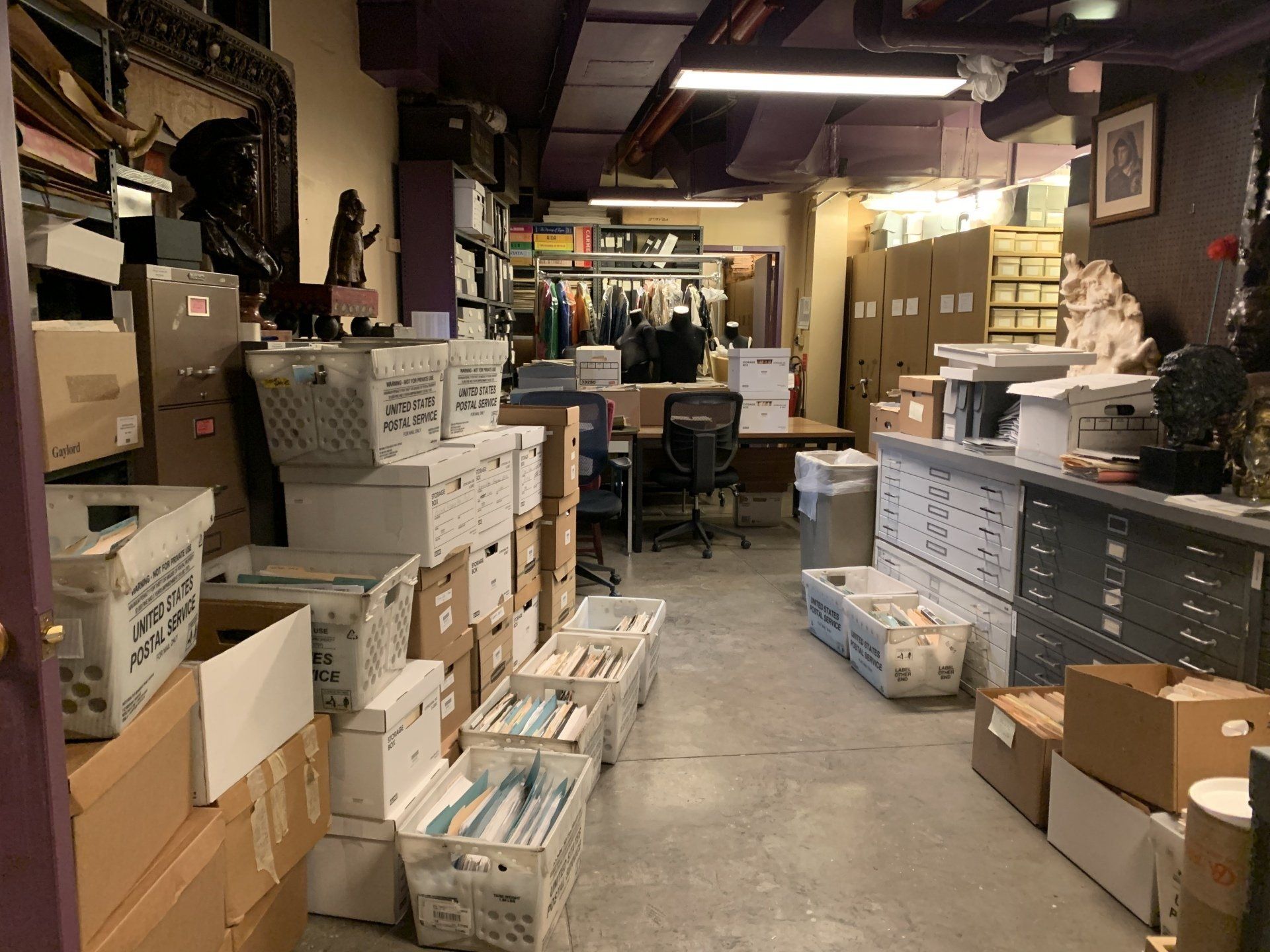 A cluttered office with boxes, files, and a desk. Fluorescent lights.