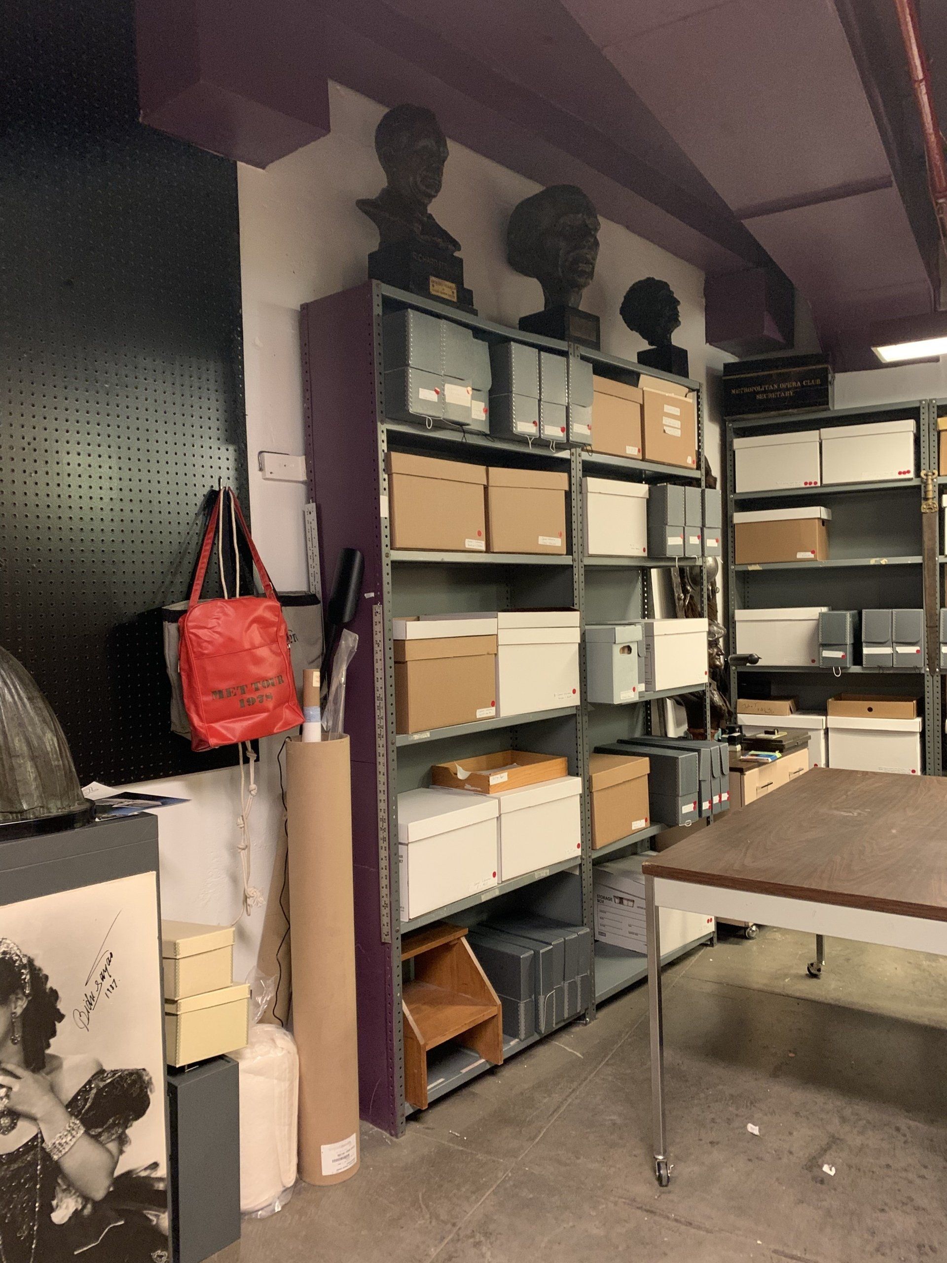 Shelves of boxes and busts in storage room; red bag hangs on black wall.