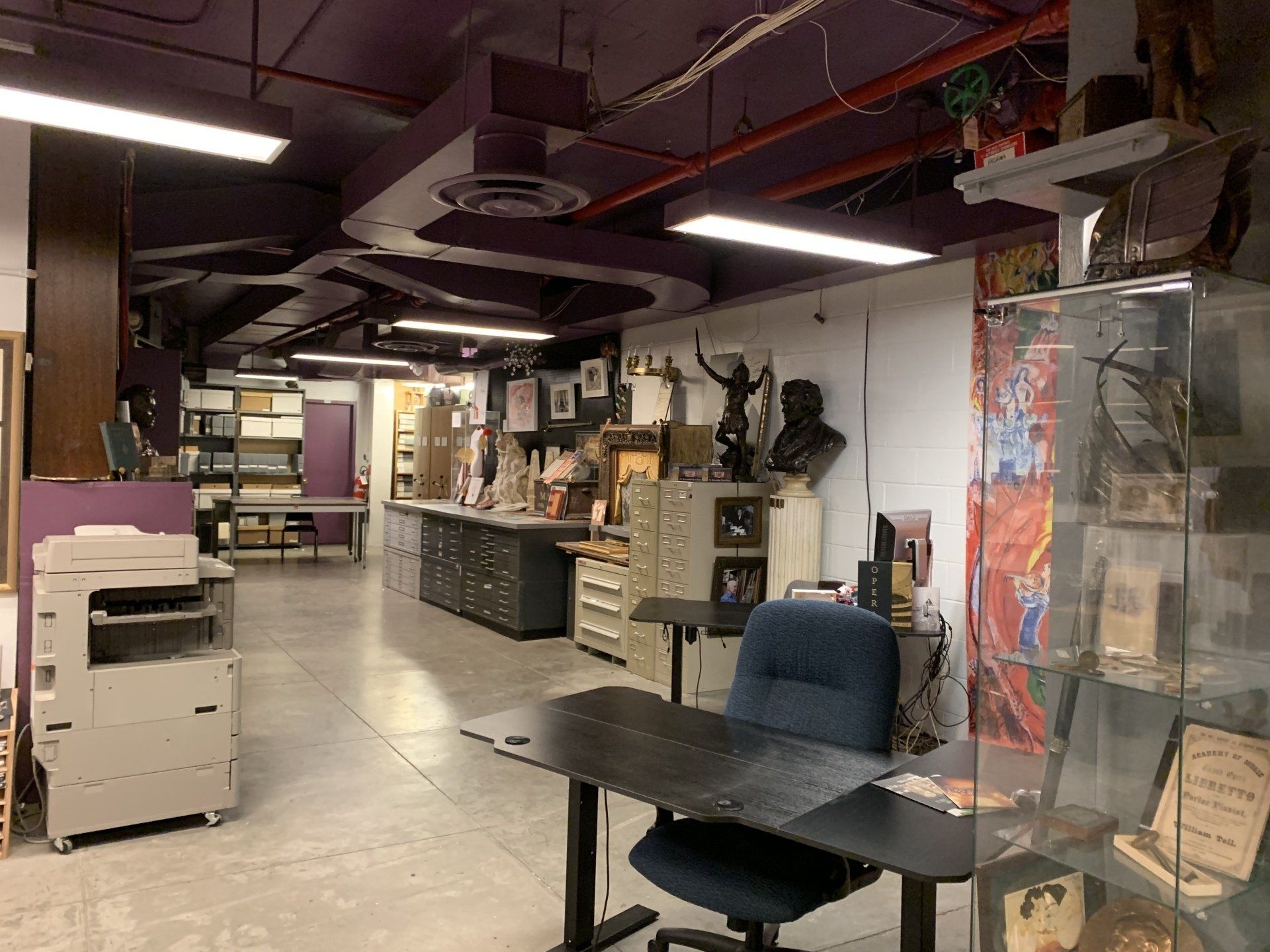 A cluttered art studio with desks, filing cabinets, sculptures, and a copier. Purple ceiling.