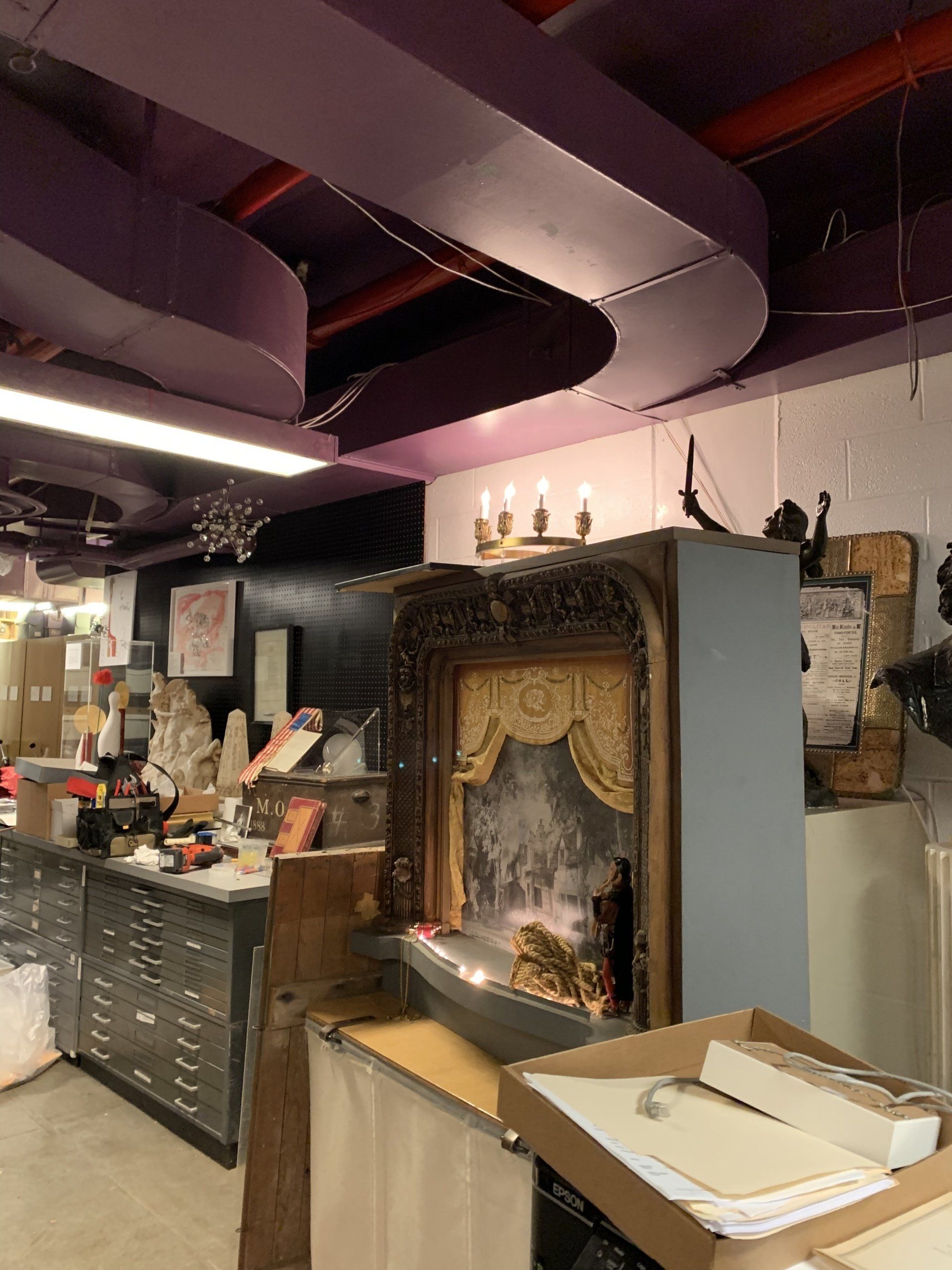 Workshop interior: dusty, with ornate stage prop, metal cabinets, purple ductwork, and dim lighting.