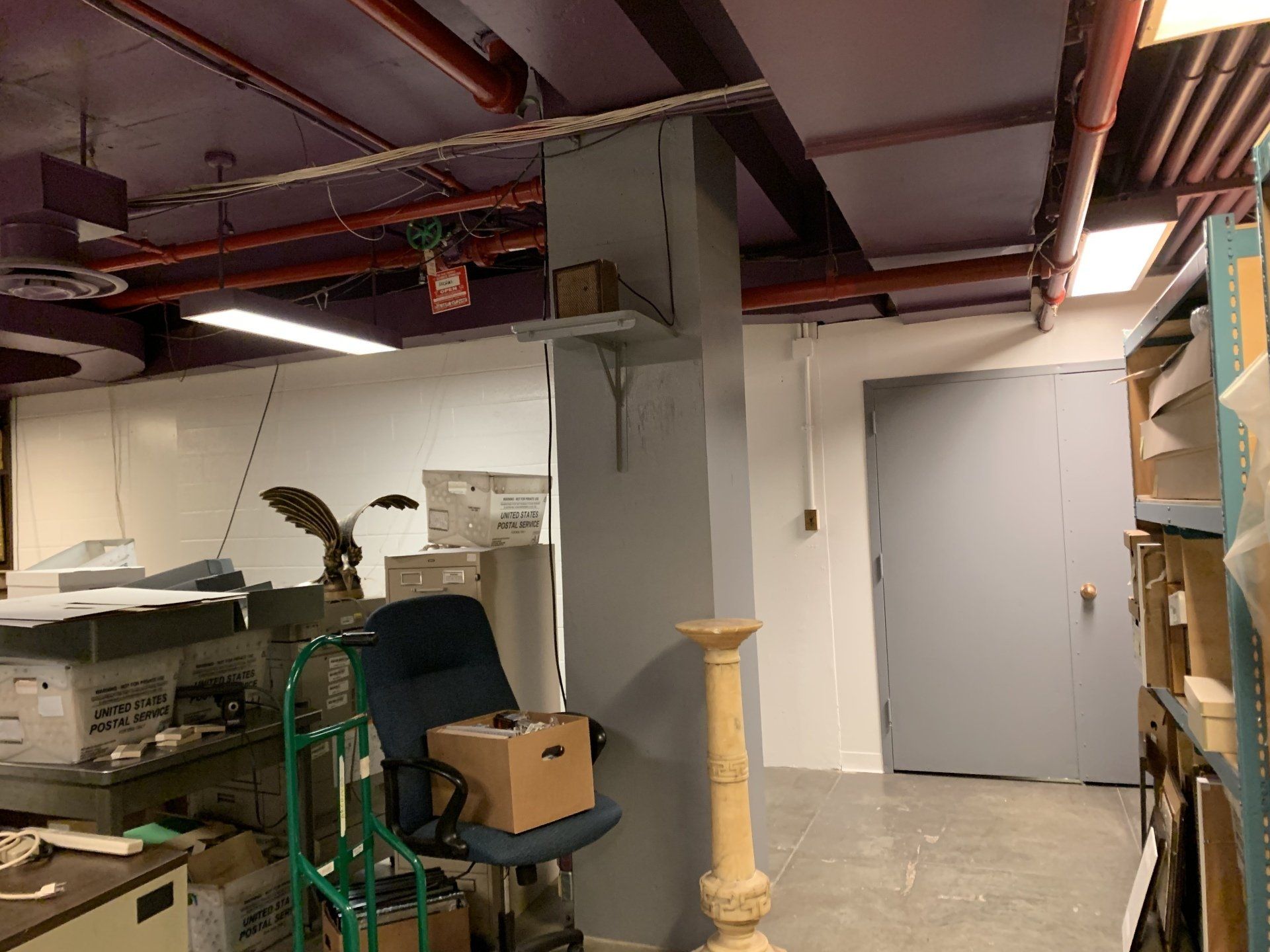 A cluttered storage room with a column, desk, chair, shelves, and a closed gray door.