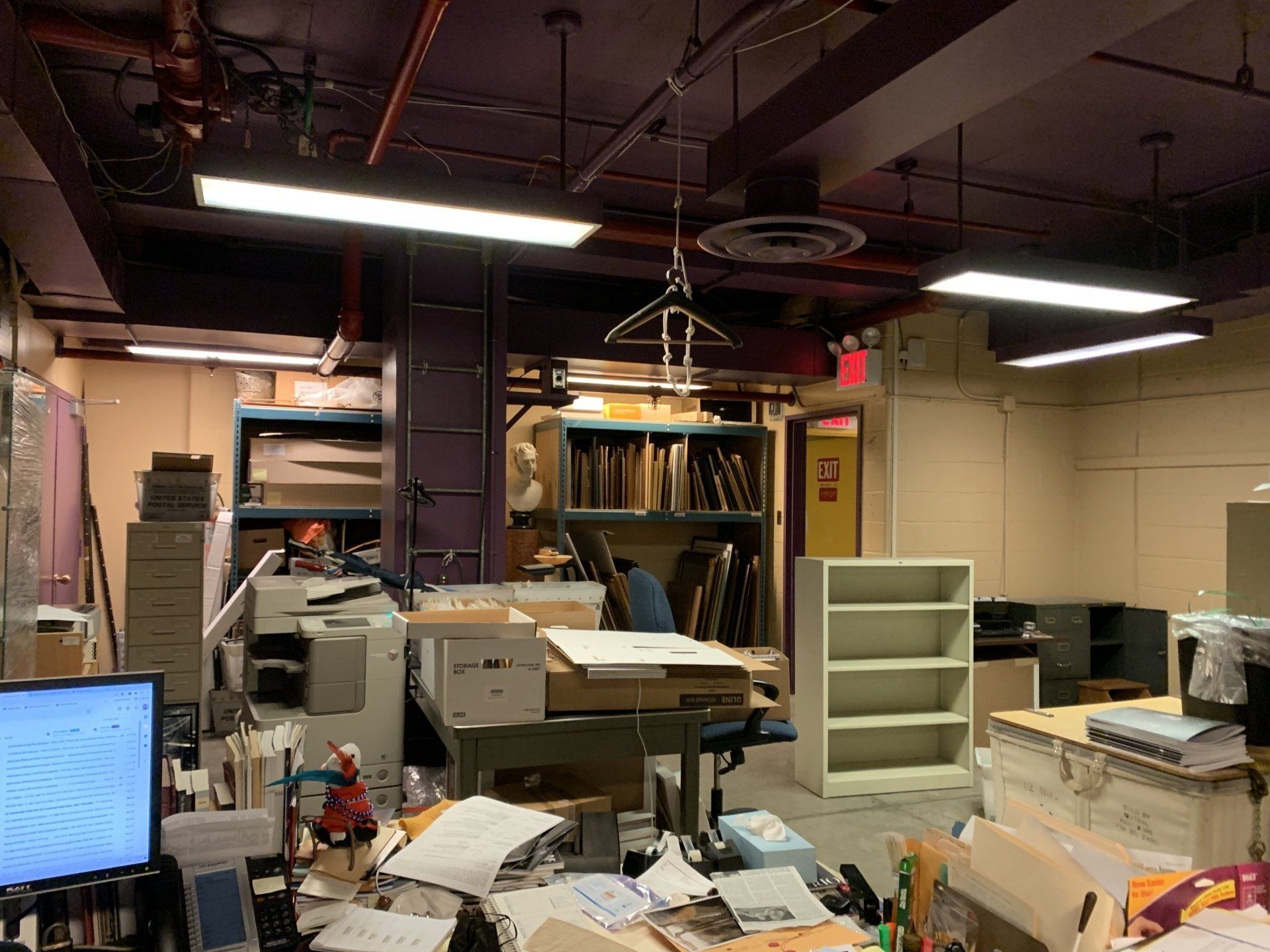 Cluttered office with purple ceiling, shelves, printer, desk, and papers.