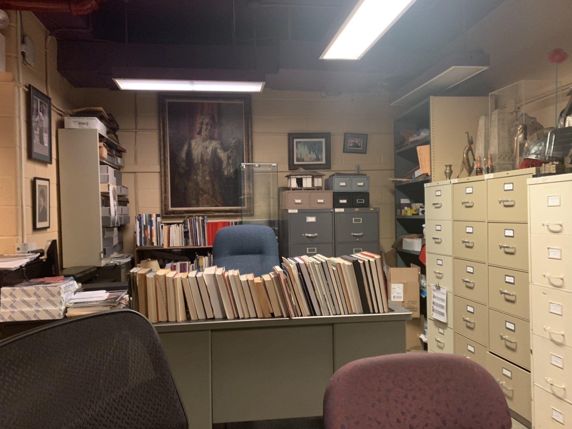 Cluttered office with books on a desk, filing cabinets, framed artwork, and an empty chair.
