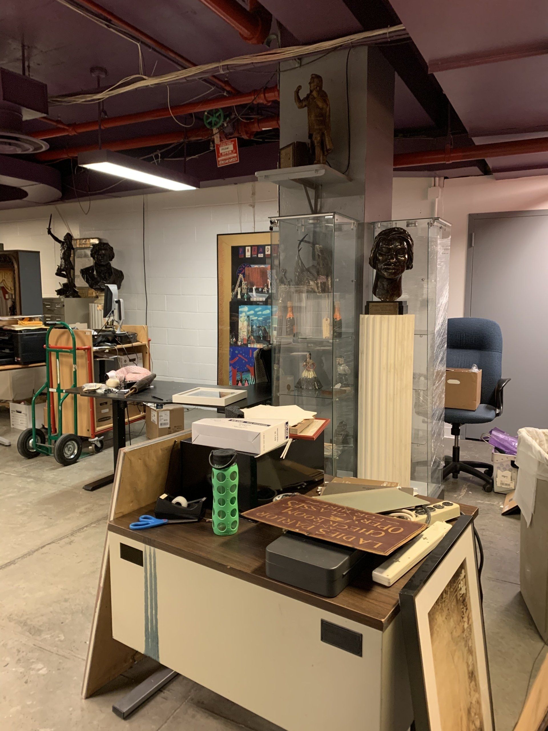 Cluttered basement room with art supplies, sculptures, and office furniture.