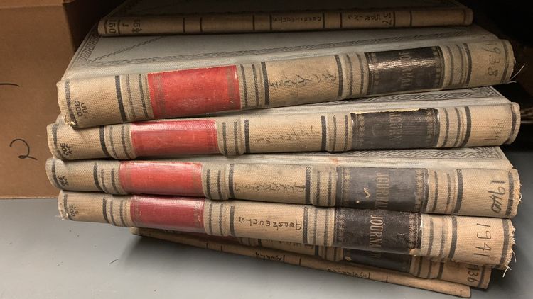 Stack of old, worn books with red and black spines, resting in a shelf.