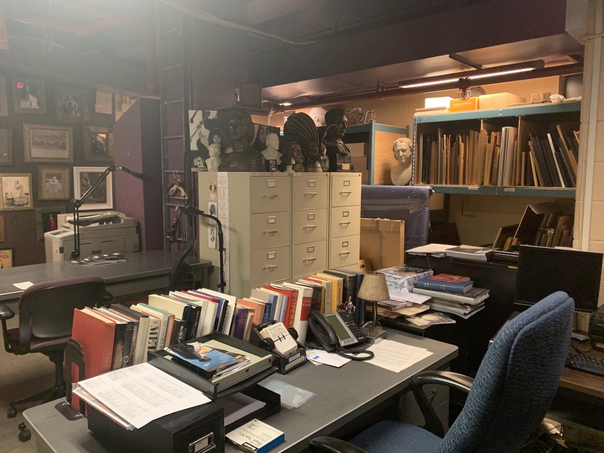 Cluttered office with desks, filing cabinets, busts, and artwork in storage.