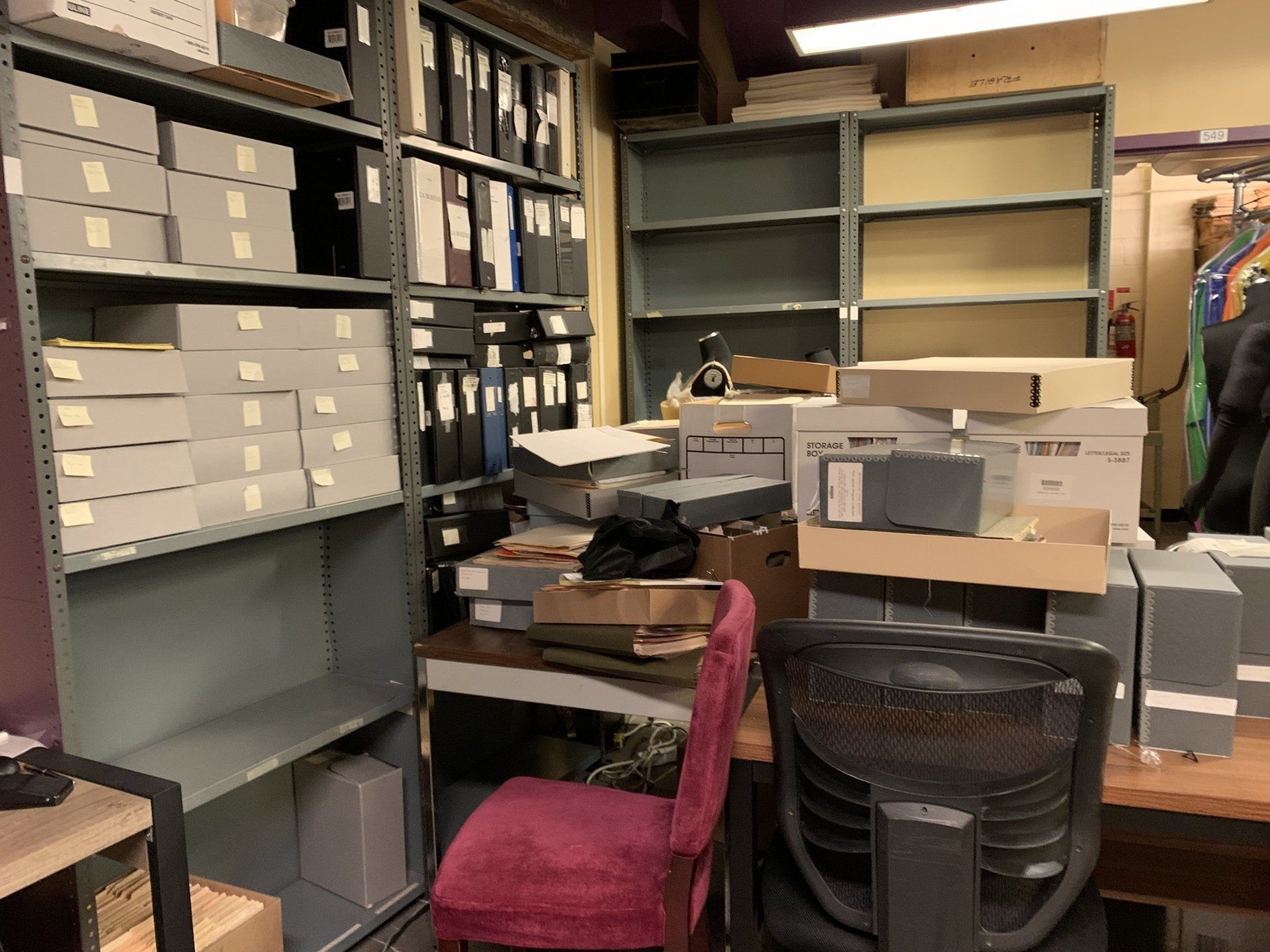 Office with shelves, boxes, files, and desk with a red chair. Overcrowded and cluttered workspace.
