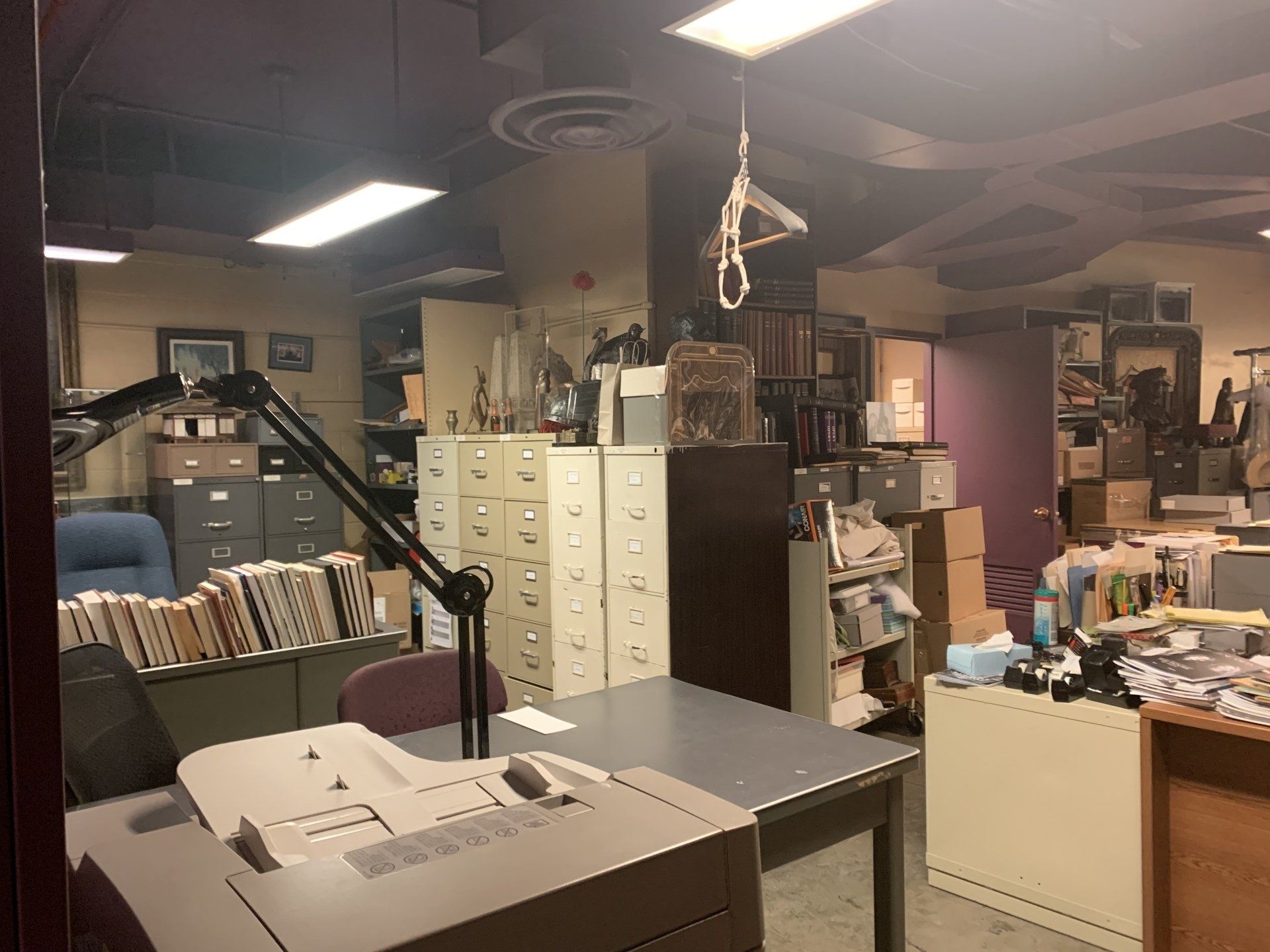Cluttered office with file cabinets, boxes, and a desk, likely a basement. Fluorescent lights.