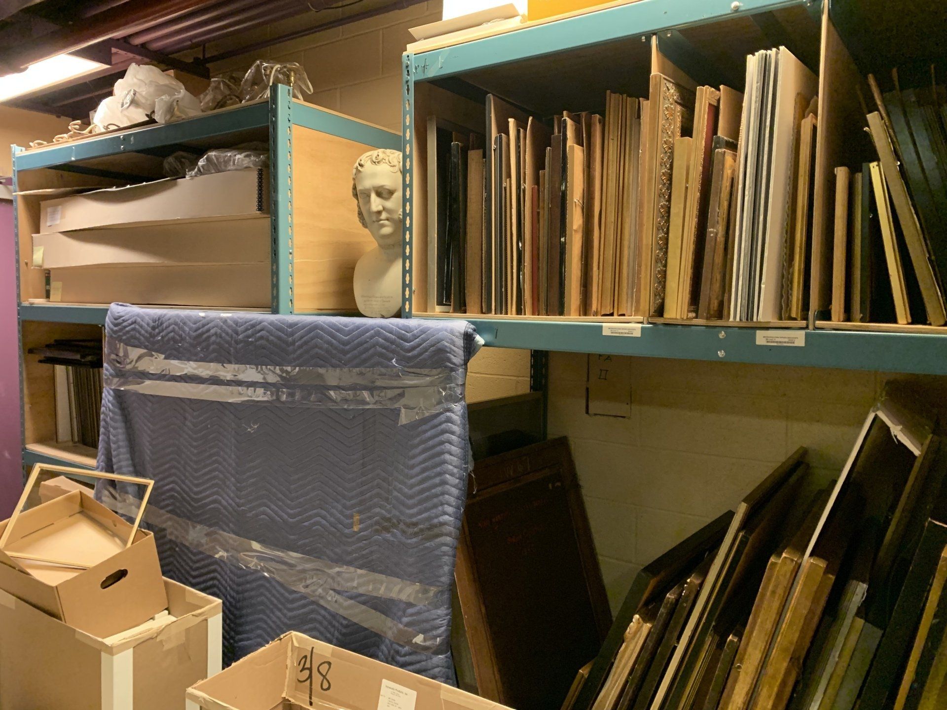 Shelves in a storage room holding artwork, frames, and busts. Boxes sit on the floor.