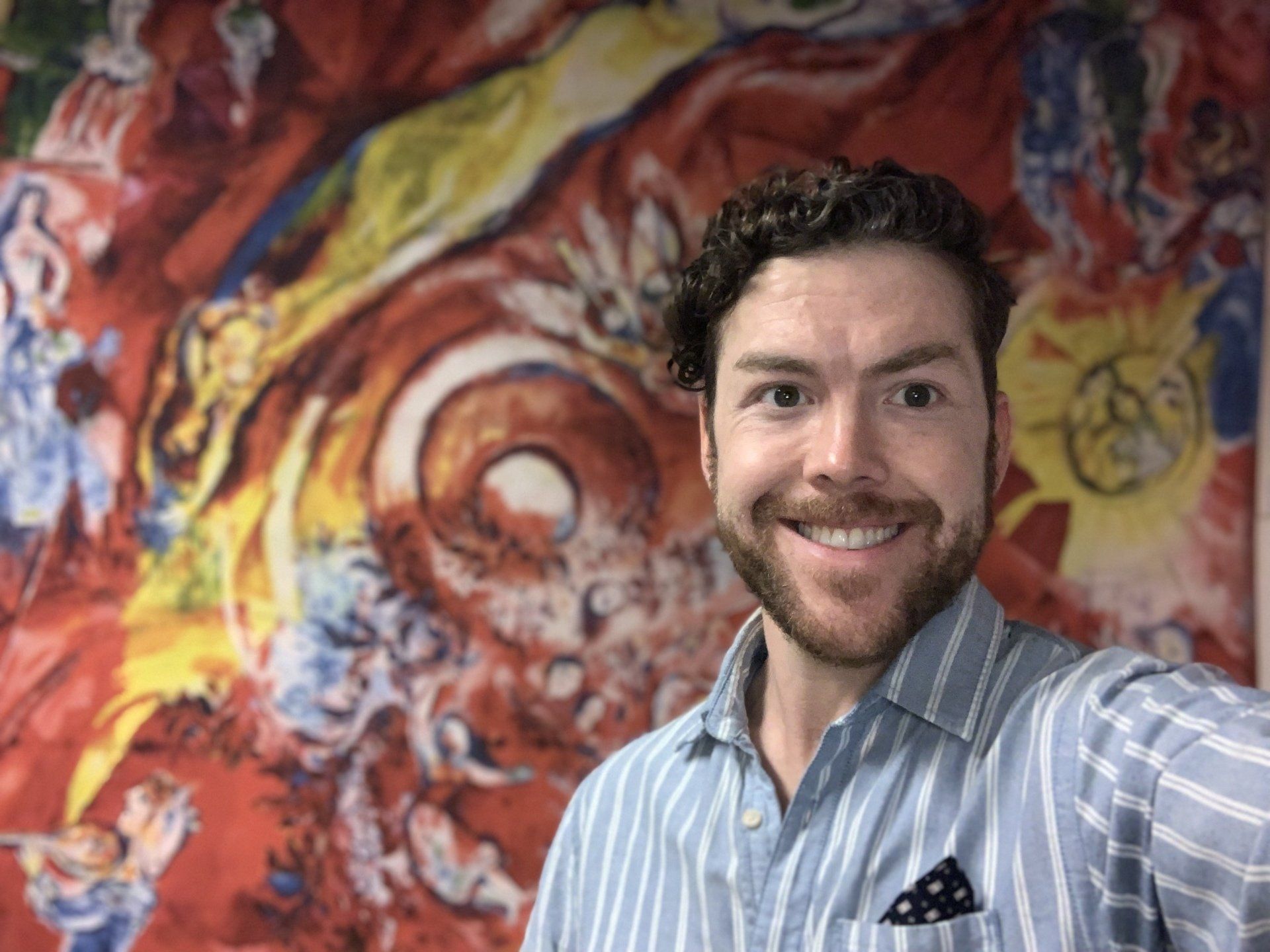 Man smiles in front of colorful abstract artwork with reds, blues, and yellows.