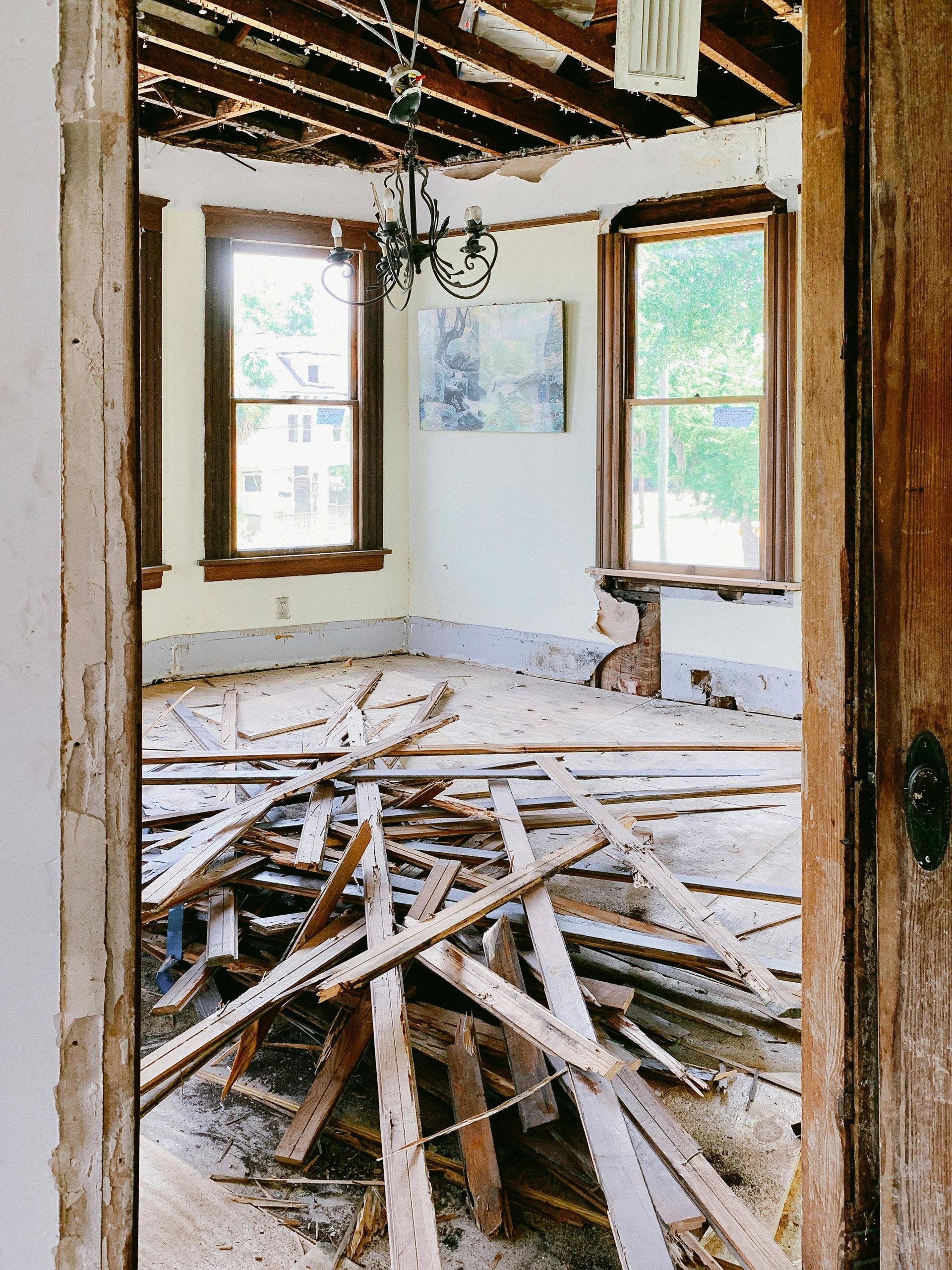 Pièce en rénovation, débris de bois au sol, poutres apparentes au plafond, deux fenêtres laissant entrevoir de la verdure.