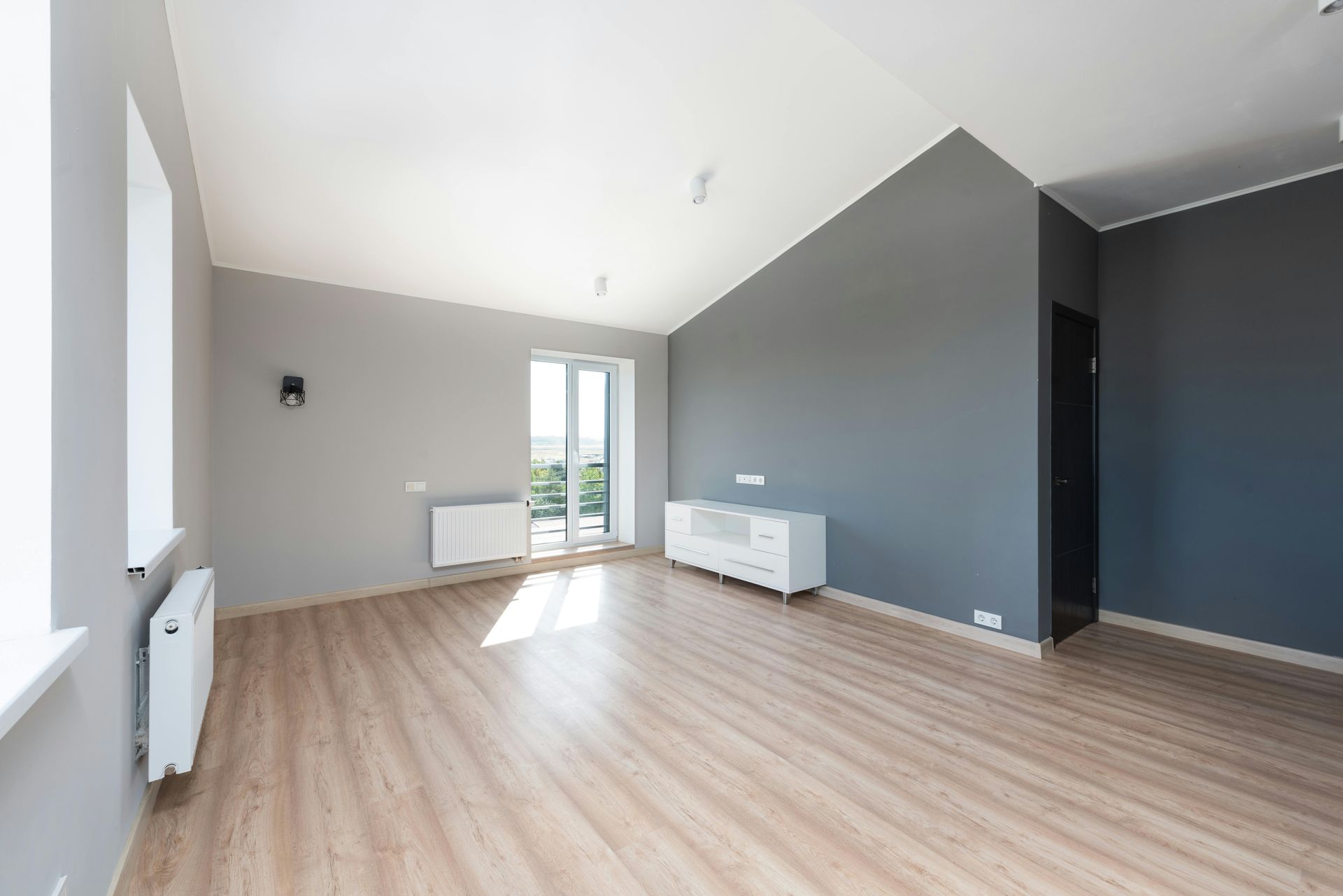 Pièce vide aux murs gris, parquet et touches de blanc, baignée de lumière naturelle par la fenêtre.