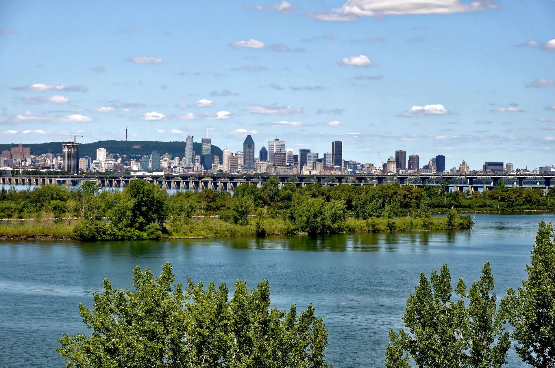 Vue de la silhouette de Montréal de l'autre côté d'une rivière, journée ensoleillée avec ciel bleu, arbres au premier plan.