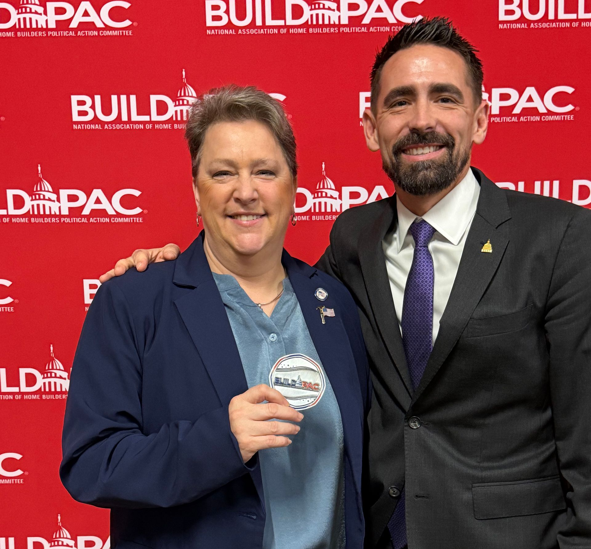 Two people pose in front of a BUILD PAC backdrop. The person on the left holds an award, smiling. The other has an arm around them, smiling.