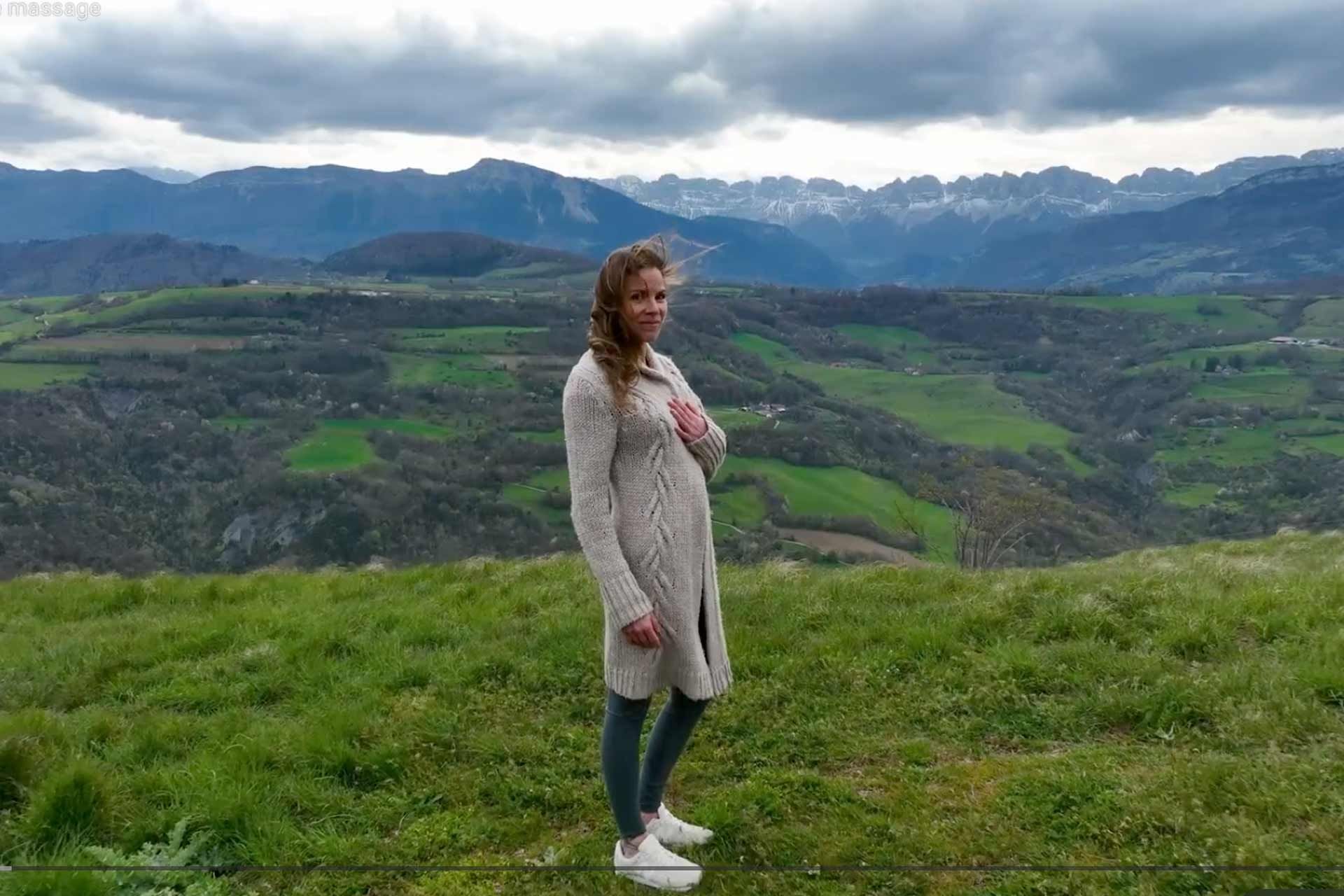 Woman in long sweater standing on grassy hill, mountain background.