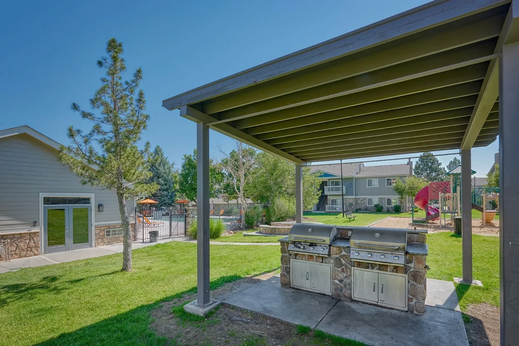Covered outdoor grill station beside a grassy yard with a house and trees in the background