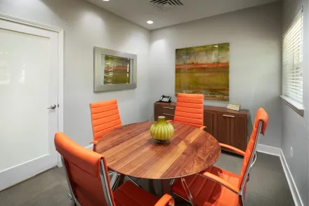 Small conference room with round wooden table, orange chairs, wall art, and a white door