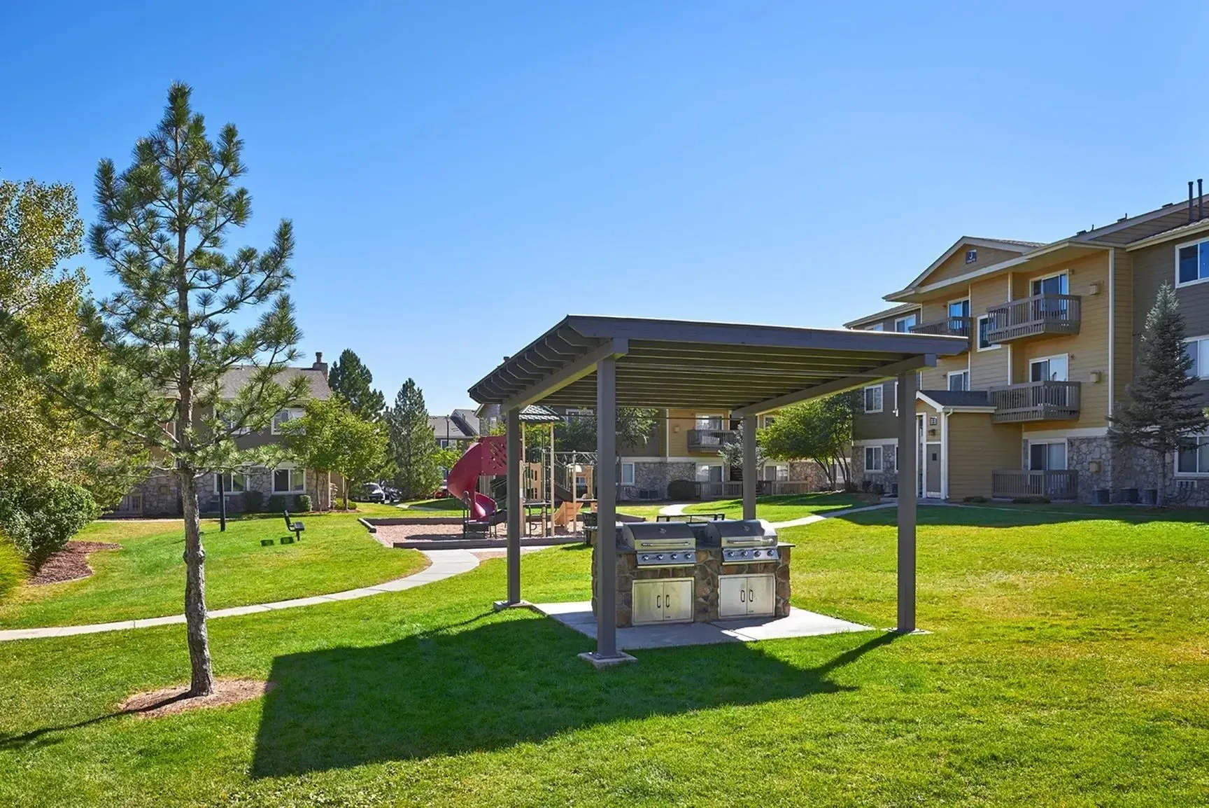 Outdoor grilling area with grills and a playground in the background at a multifamily property.
