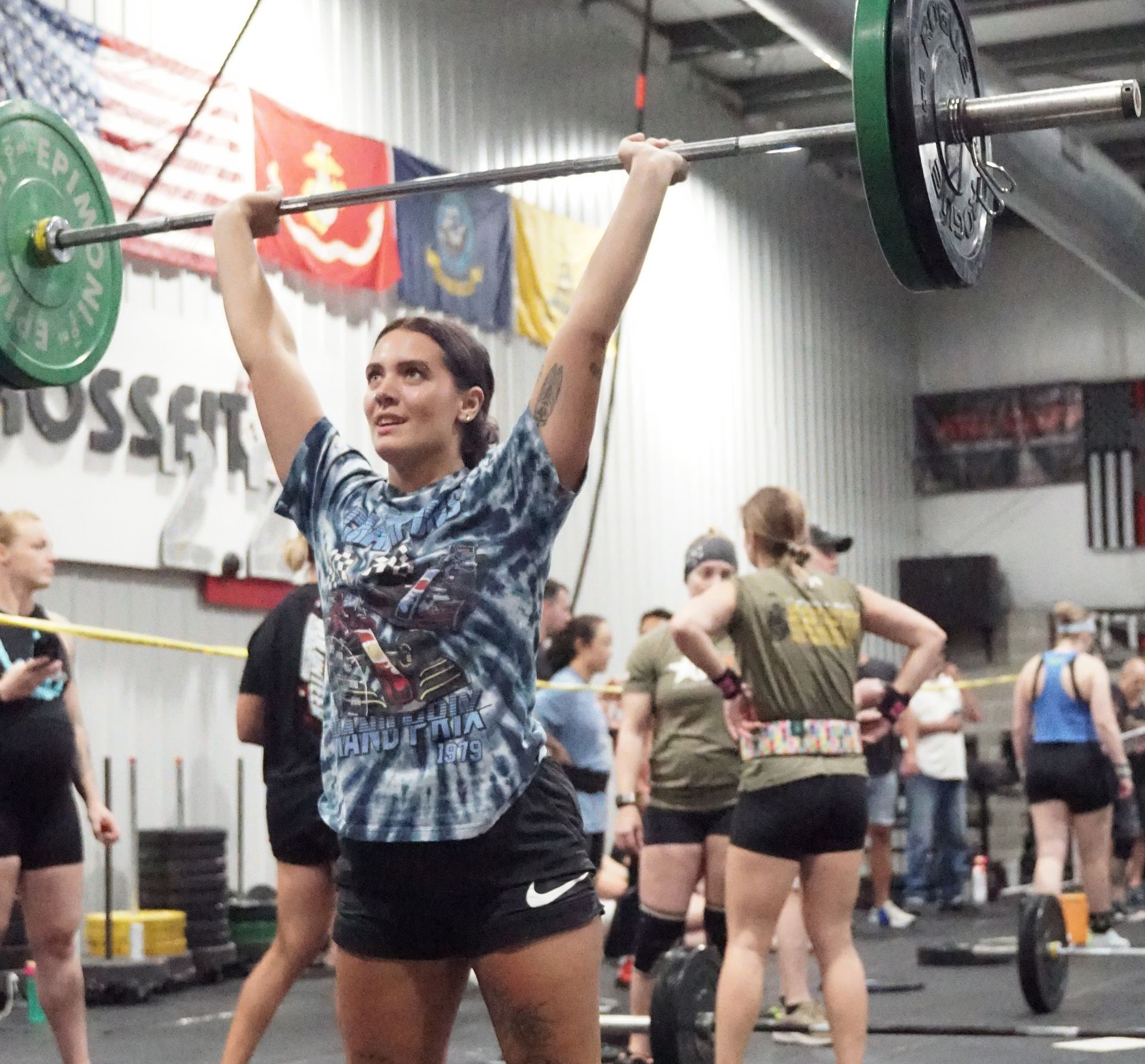 Woman in black tank top and maroon leggings using an air bike in a gym, another person watches.