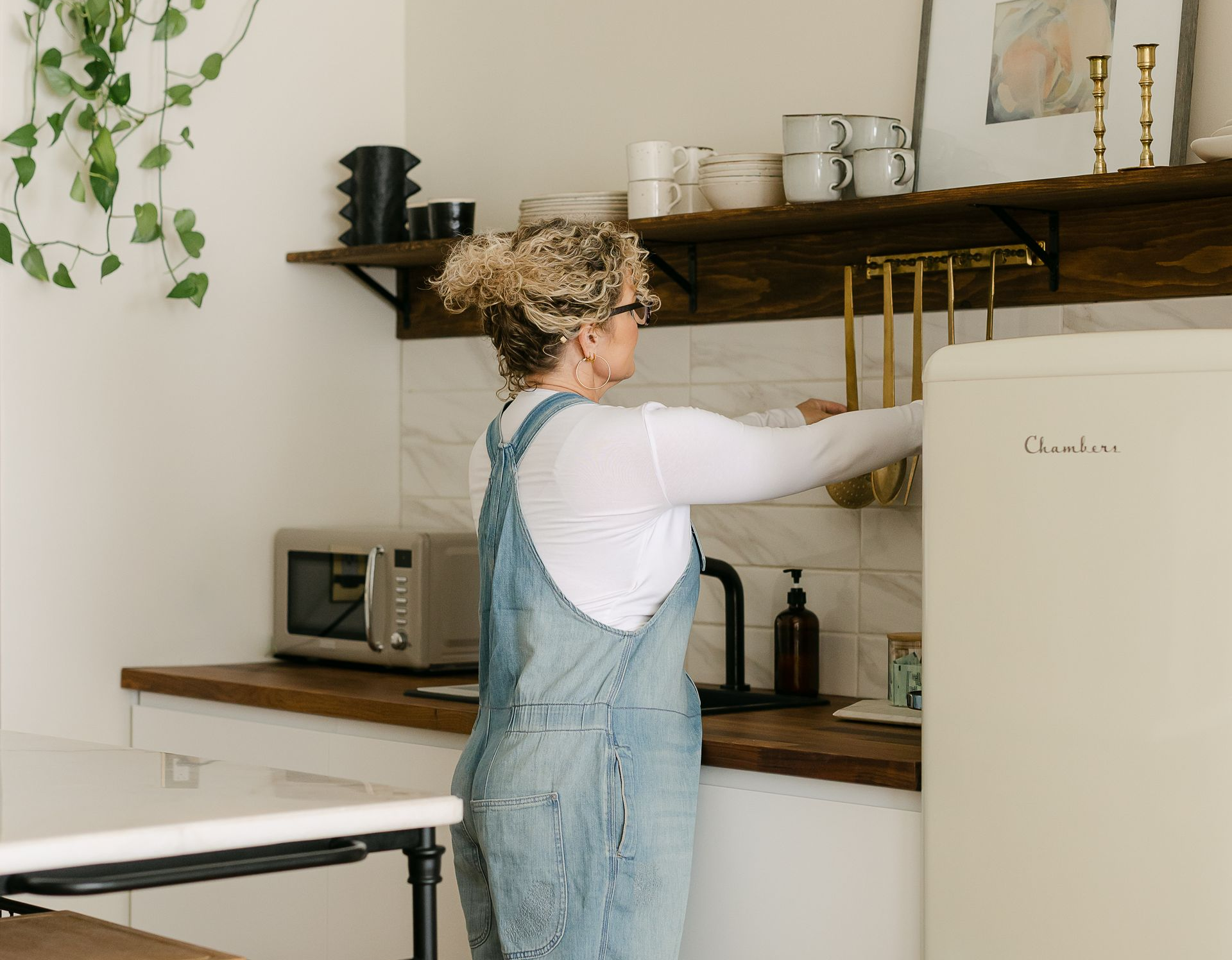 Woman in denim overalls in a kitchen reaching for hanging utensils near a cream-colored refrigerator.