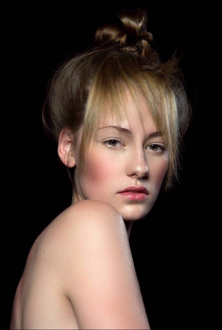 Ritratto di una persona con i capelli raccolti in uno chignon e una frangia sfilata, che guarda verso la fotocamera su uno sfondo nero.