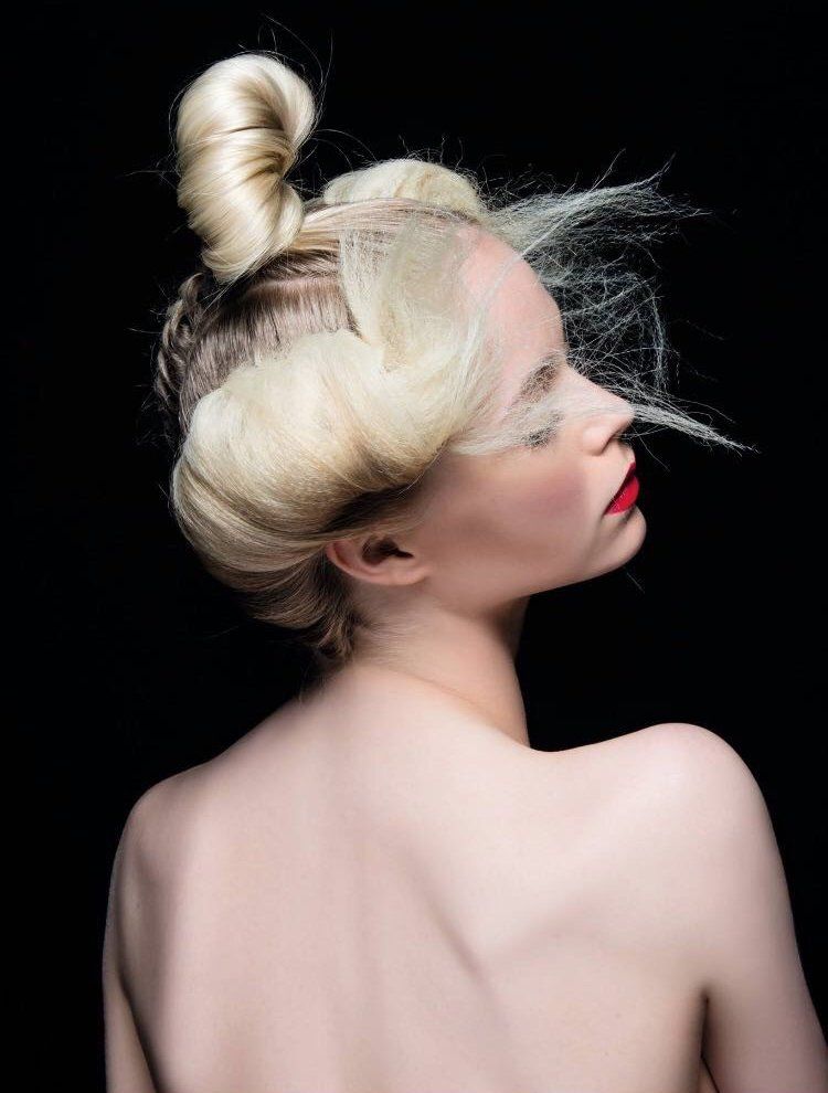 Ritratto di profilo di una persona con capelli biondo platino raccolti in uno chignon alto e morbide onde, su sfondo nero.